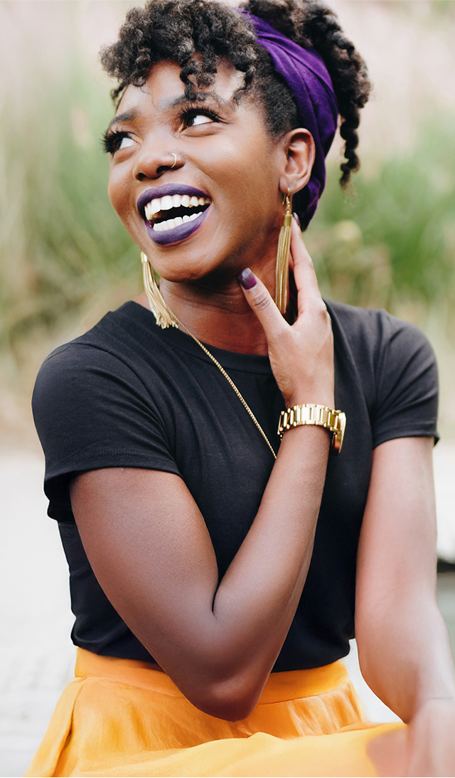 A woman smiling happily outdoors, wearing a purple headscarf, gold jewelry, and a black top with a yellow skirt.