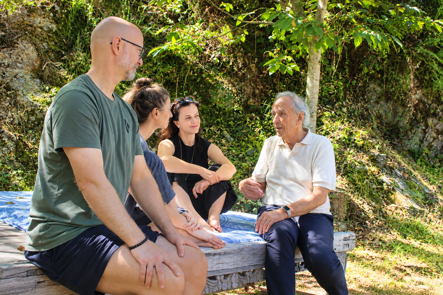 Five people sitting on a wooden platform outdoors, engaging in a conversation, with a green, leafy background.
