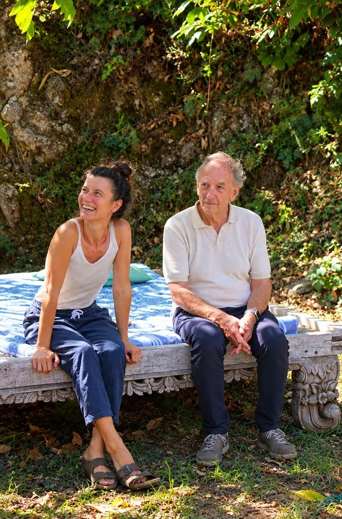 A woman and an older man sitting on an ornate wooden bench outdoors surrounded by green foliage and rocks, enjoying a sunny day.