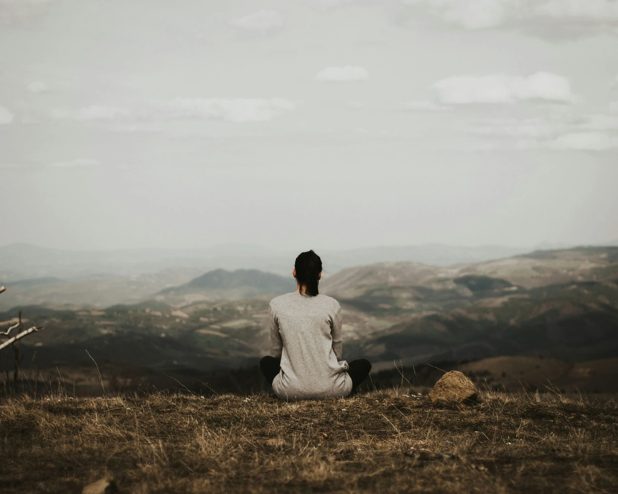 A person sitting cross-legged on a grassy hilltop, facing a vast landscape of rolling hills and mountains under a cloudy sky.