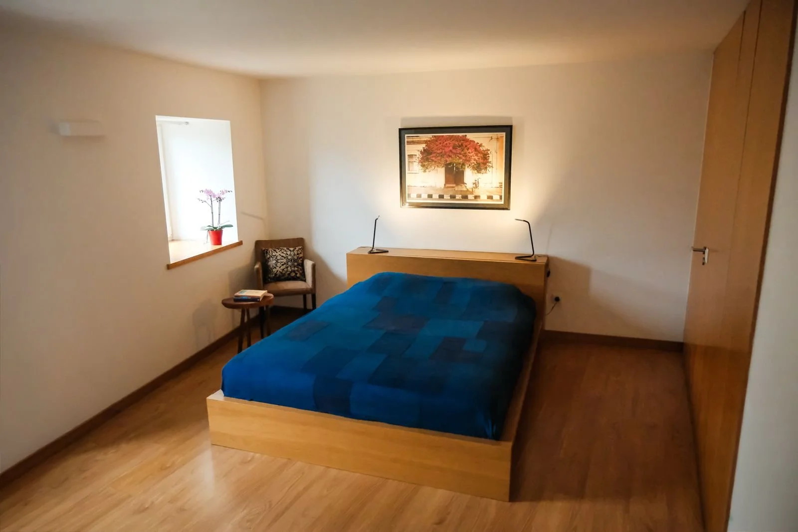 A bedroom with a wooden bed frame and blue bedding, a small bedside table, an armchair with a patterned cushion, a window with a potted orchid, and a framed picture on the wall.