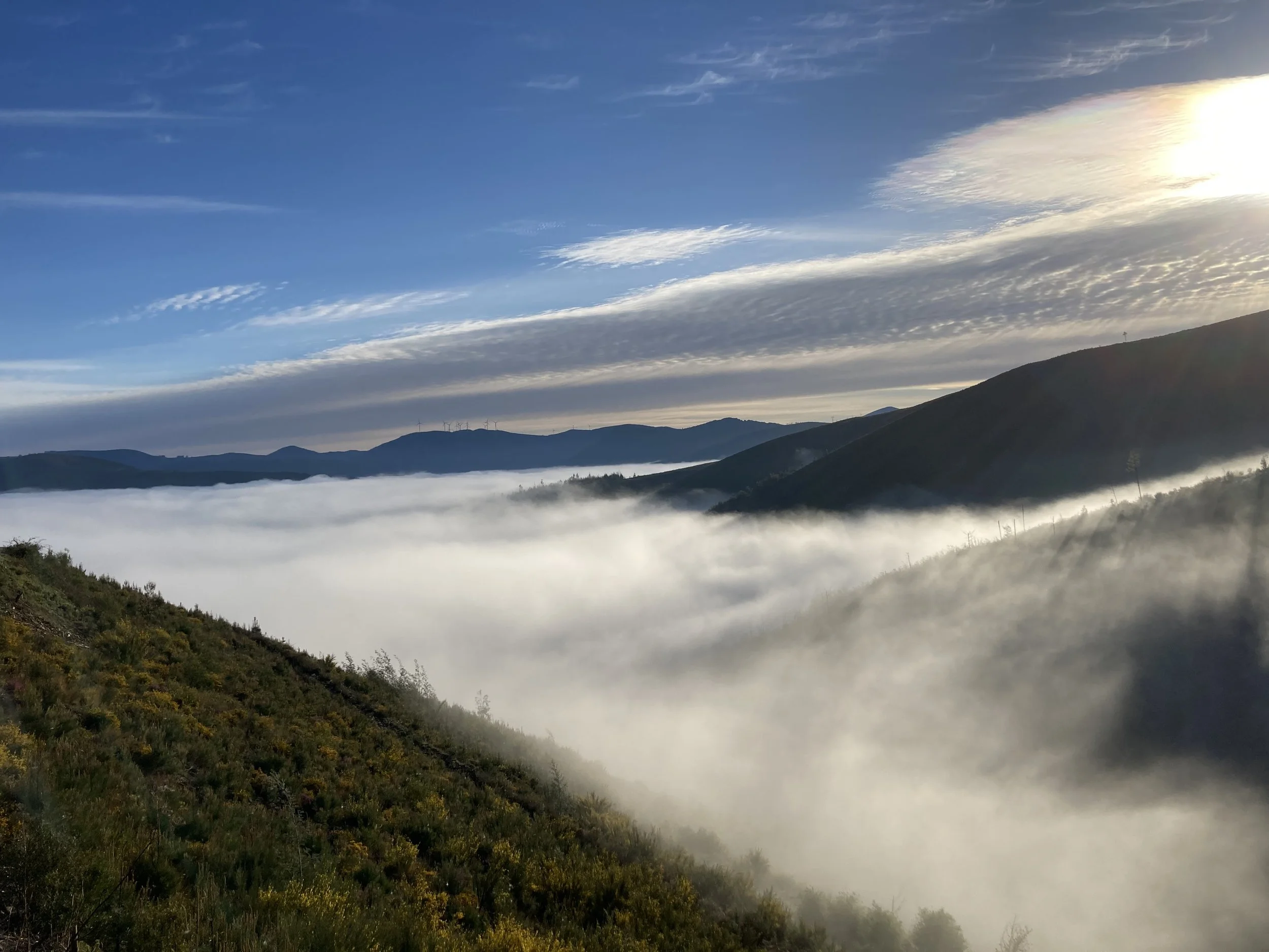Mountain landscape with a cloudy sky, mist hovering over the hills, and wind turbines visible on the distant ridges during sunrise or sunset.