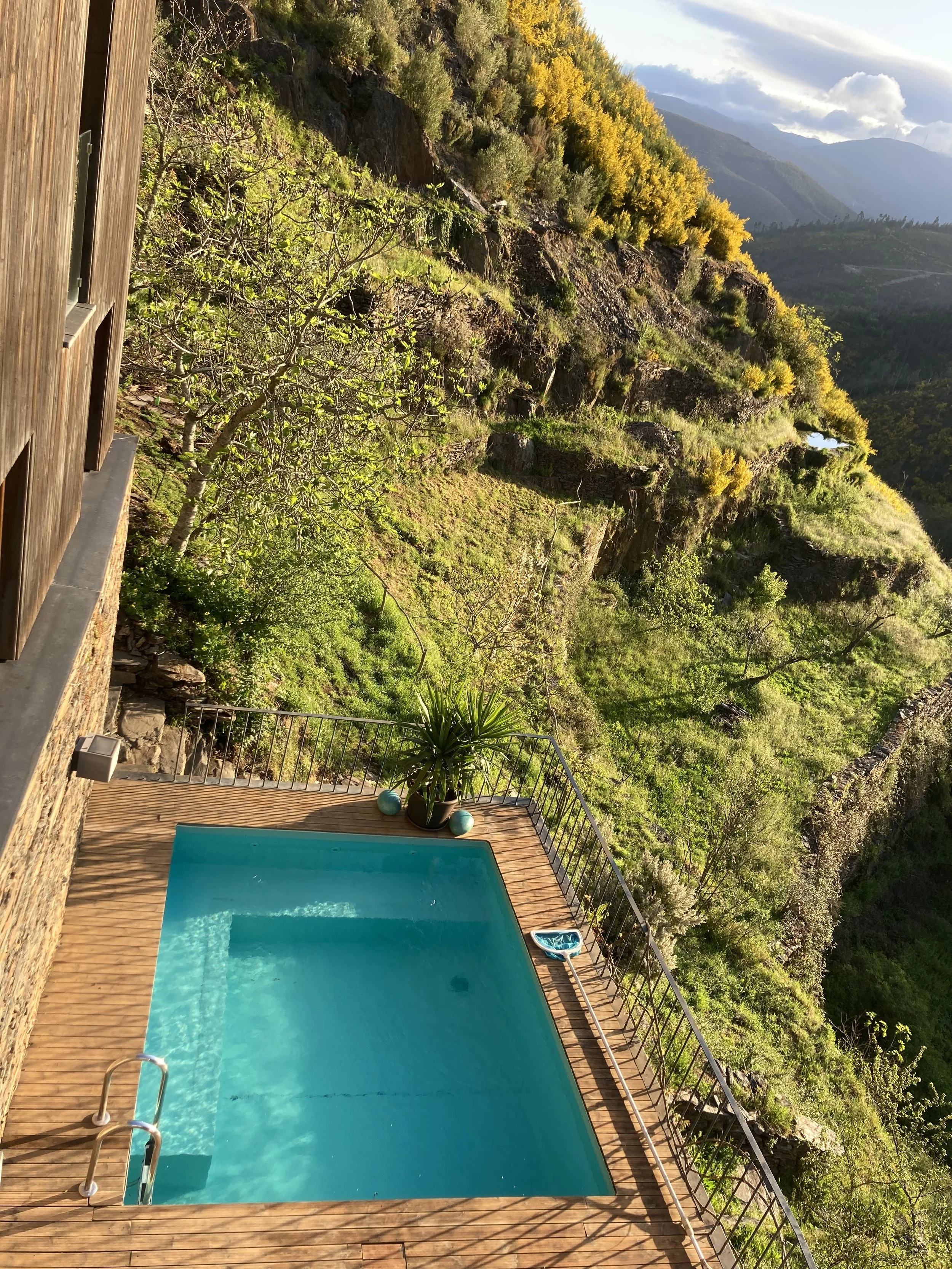 A small outdoor swimming pool on a wooden deck, surrounded by a metal railing, with a potted plant and decorative balls, overlooking a hillside with trees and mountains in the background during daytime.