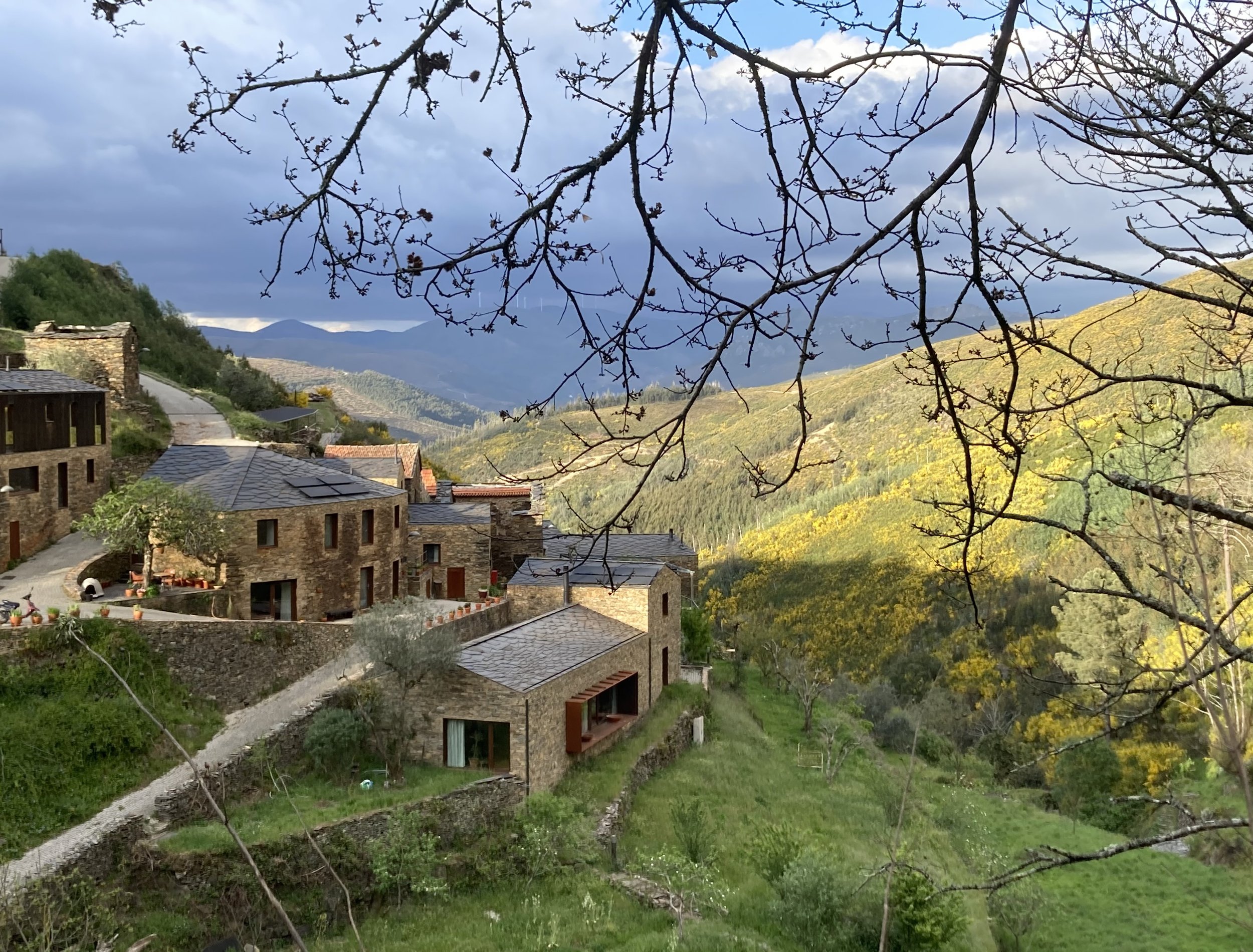 A scenic view of a hillside with stone houses and a winding road, surrounded by green trees and mountains in the background, under a cloudy sky.