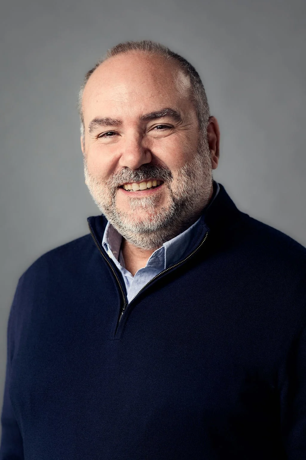 Headshot of a smiling middle-aged man with a beard, wearing a navy sweater over a collared shirt, against a neutral grey background.