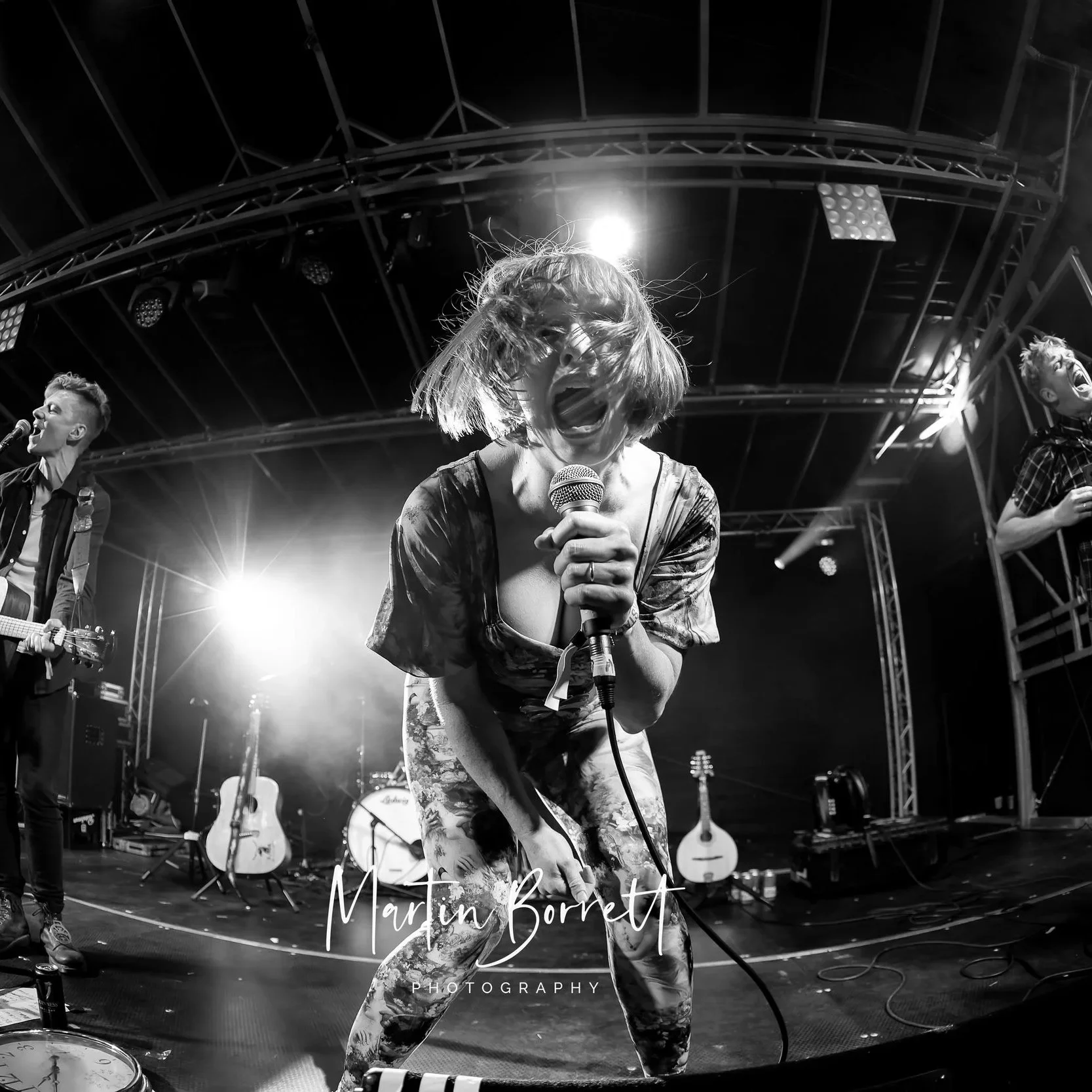 Black and white photograph of a woman passionately singing into a microphone on stage with her hair covering her face. Two men are visible in the background, one playing guitar and the other also singing, with stage lights shining behind them.