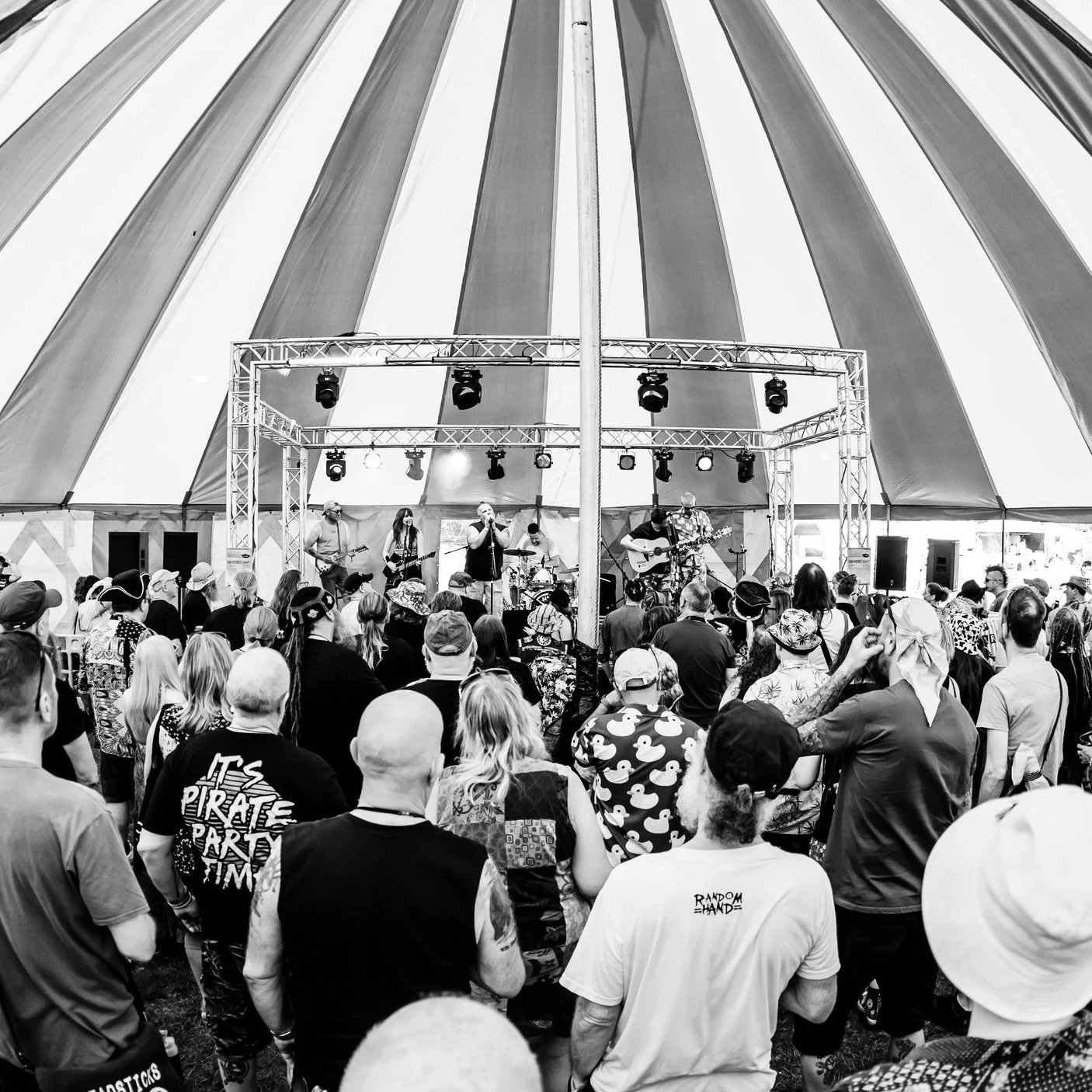 A black and white photo of a live music performance inside a large striped tent with an audience watching the band.