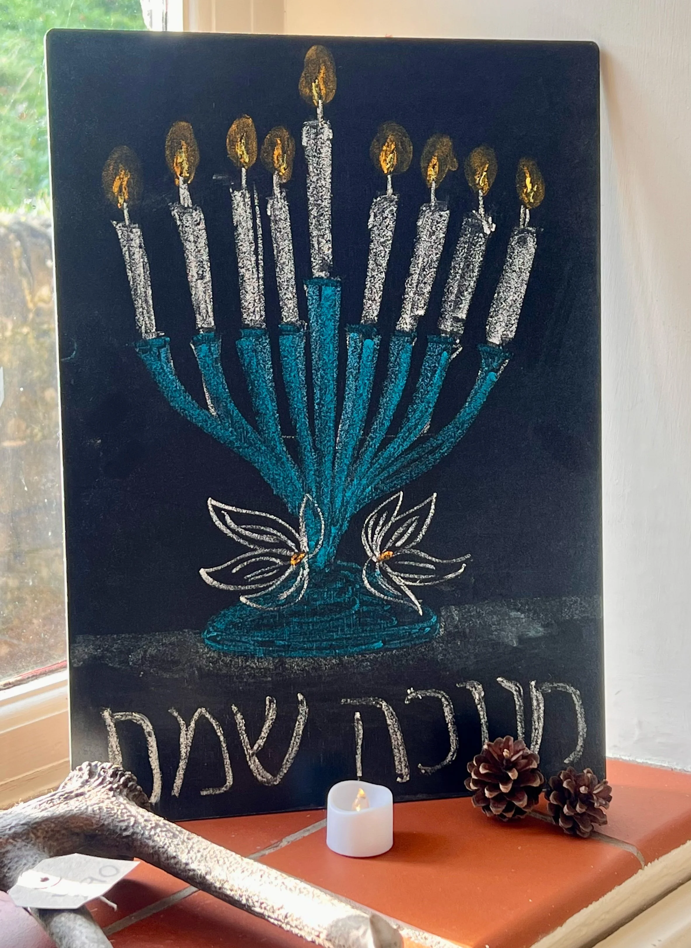 A blackboard with a colorful drawing of a menorah with candles, two white flowers, Hebrew text, a small white candle, and two pinecones on a windowsill.