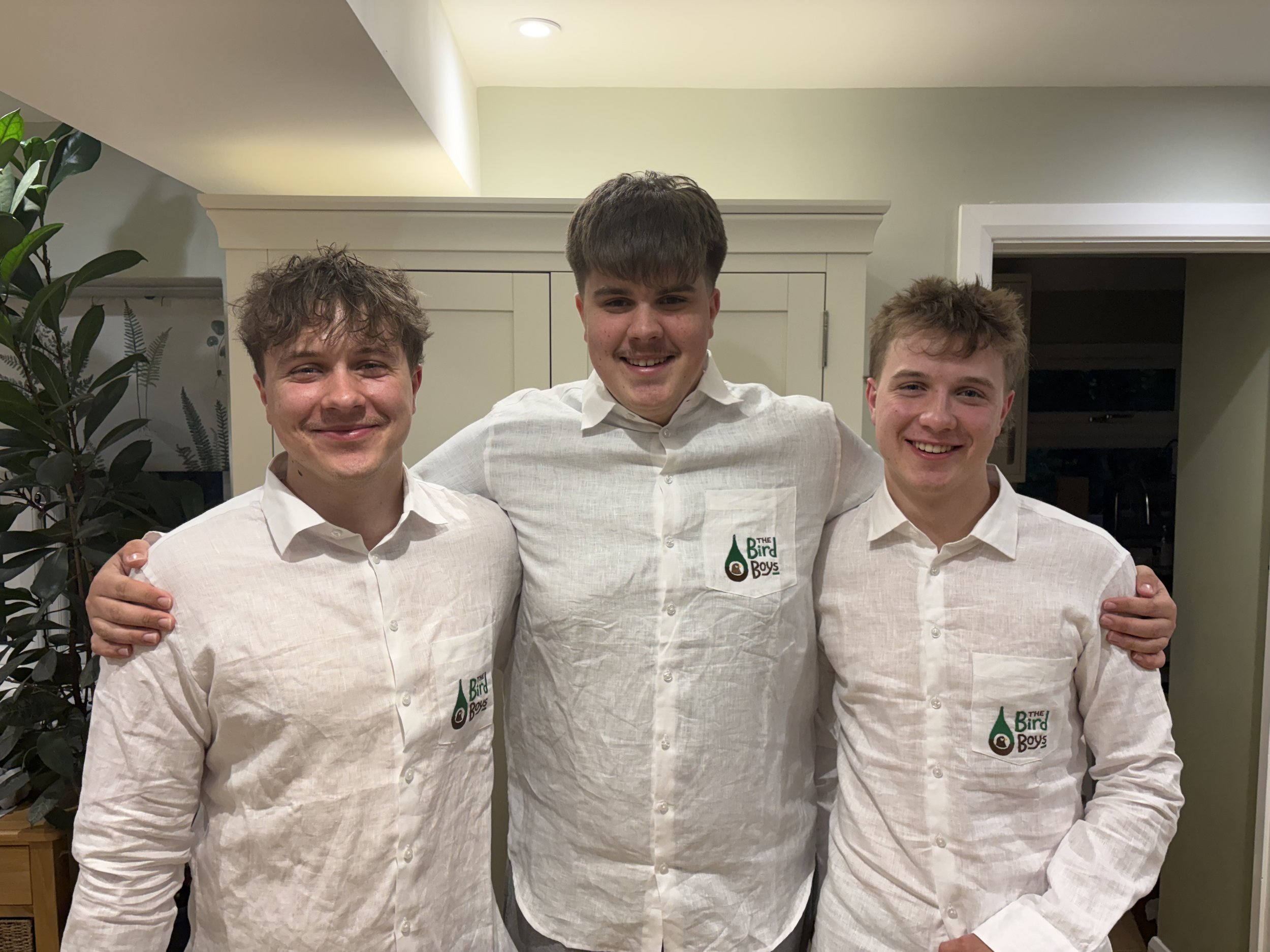Three young men wearing matching white shirts with a logo reading 'The Bird Boys' standing indoors with their arms around each other, smiling at the camera.