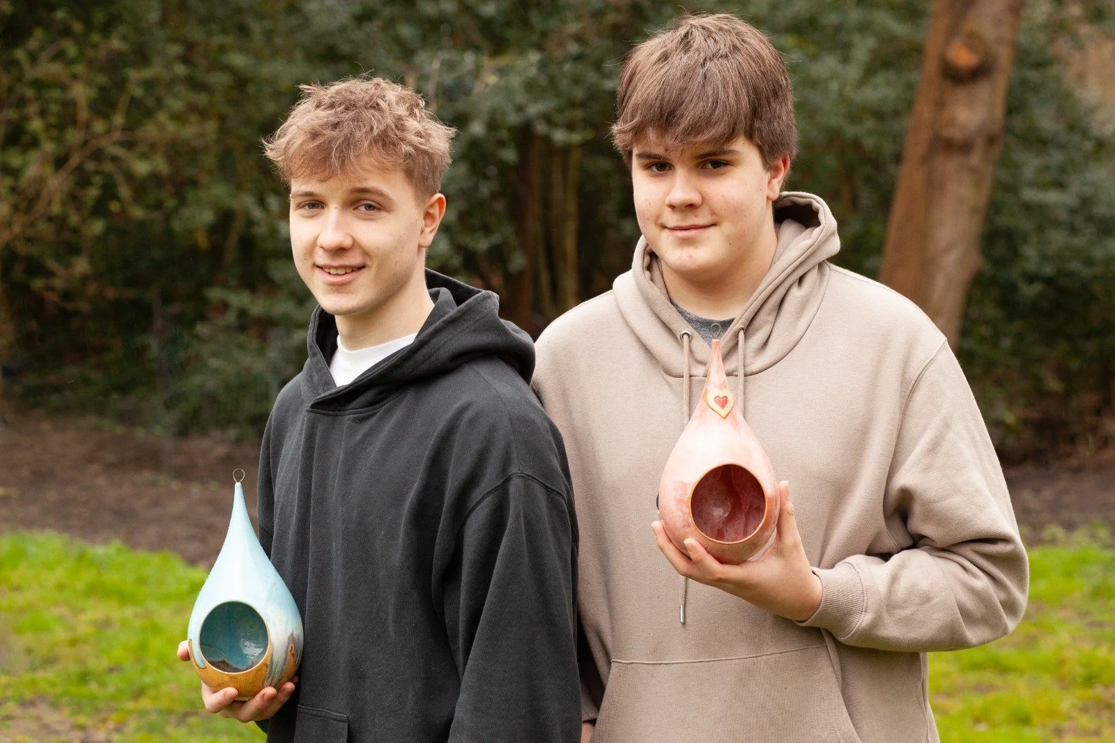 George and Zac, the bird boys  holding ceramic bird feeders, one blue and one pink, in a park with trees and grass.