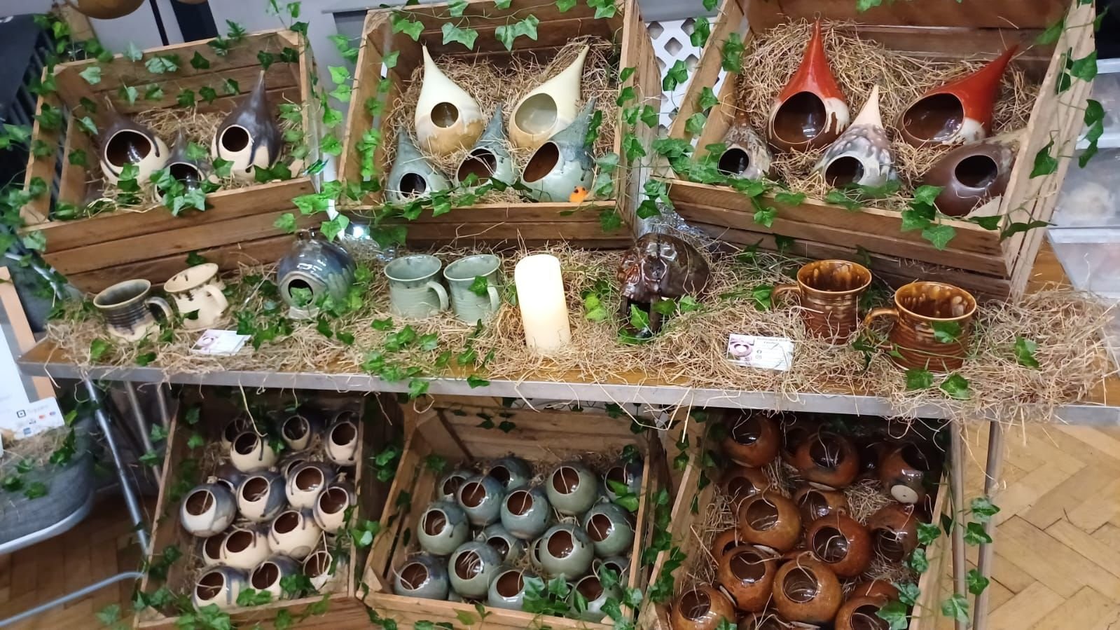 Display of handcrafted ceramic birdhouses and pottery cups arranged in wooden crates decorated with fake green vines and straw, with a candle in the center.