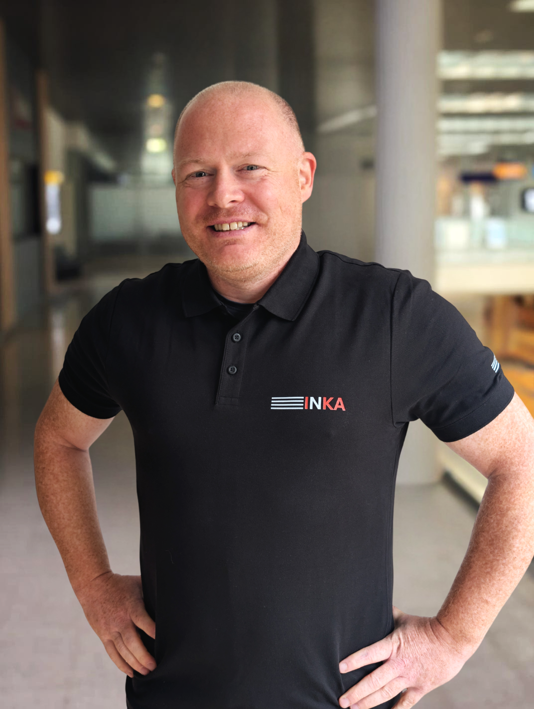 Ein Mann trägt ein schwarzes polohemd mit dem Logo 'INKA' auf der linken Brustseite, steht in einem modernen, hellen Innenraum und lächelt in die Kamera.