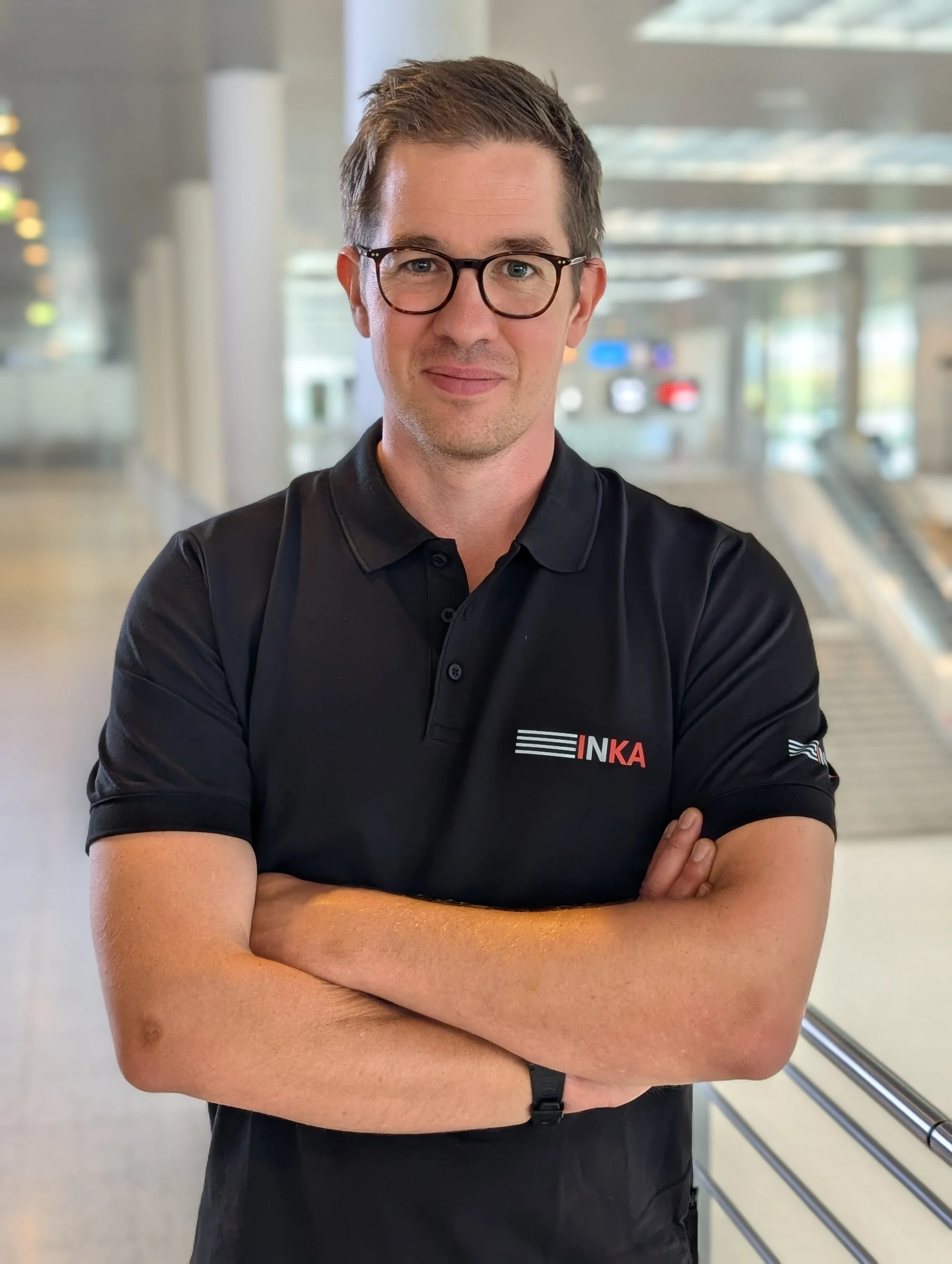 Ein Mann mit Brille steht in einer modernen, hell erleuchteten Halle. Er trägt ein schwarzes Polo-Shirt mit dem Logo 'INKA' auf der Brust, seine Arme sind verschränkt.