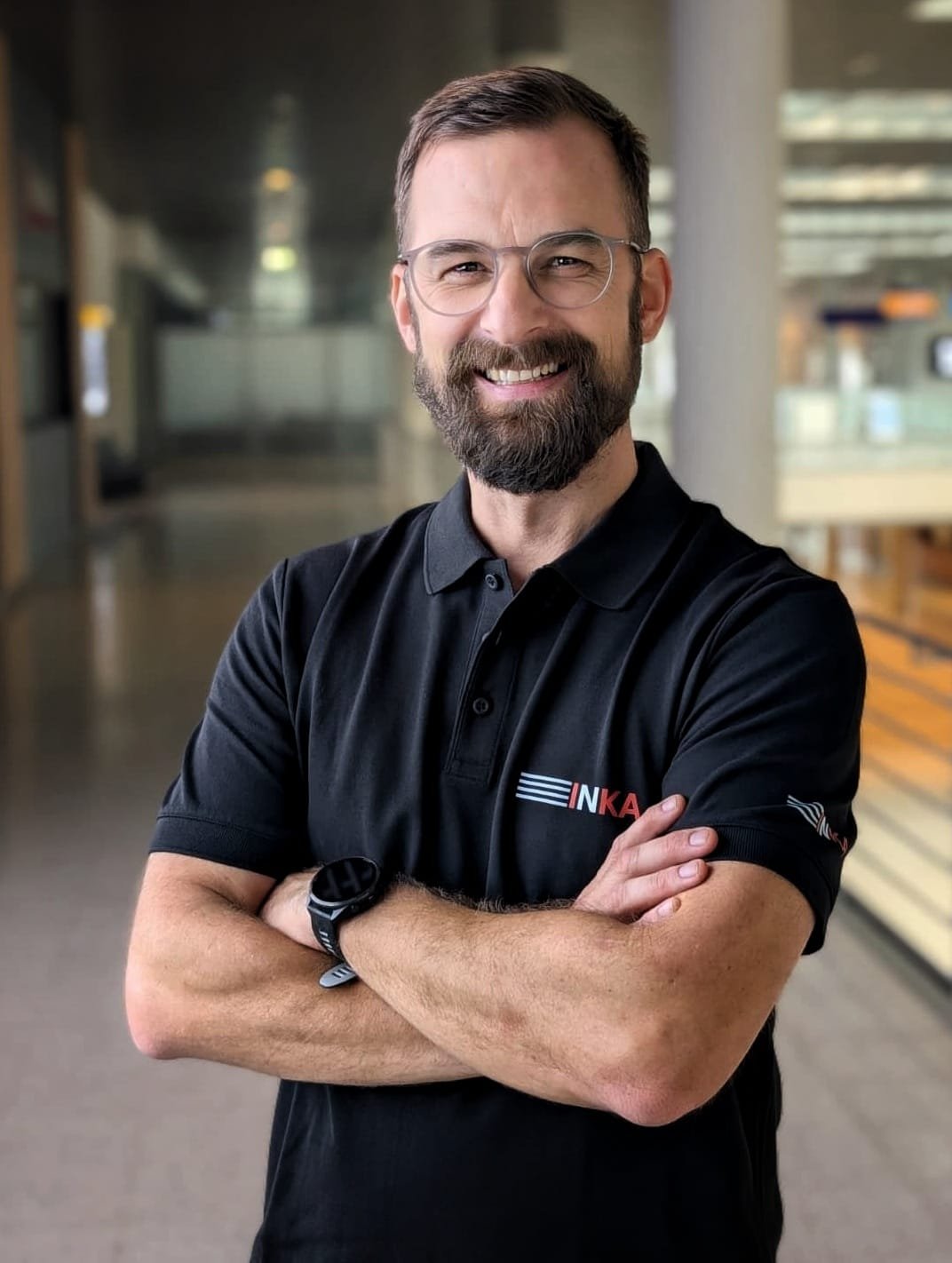 Ein lächelnder Mann mit Brille und Bart, trägt ein schwarzes Polo-Shirt mit Logo, steht in einem modernen Gebäude mit hellem Holz und Glas im Hintergrund.