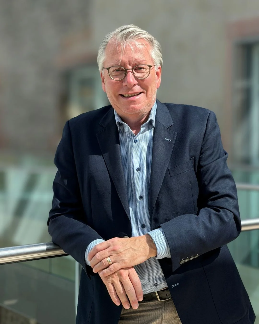 Ein Mann mit Brille in einem dunklen Blazer und blauer Hemd vor hellem Hintergrund, lächelnd.