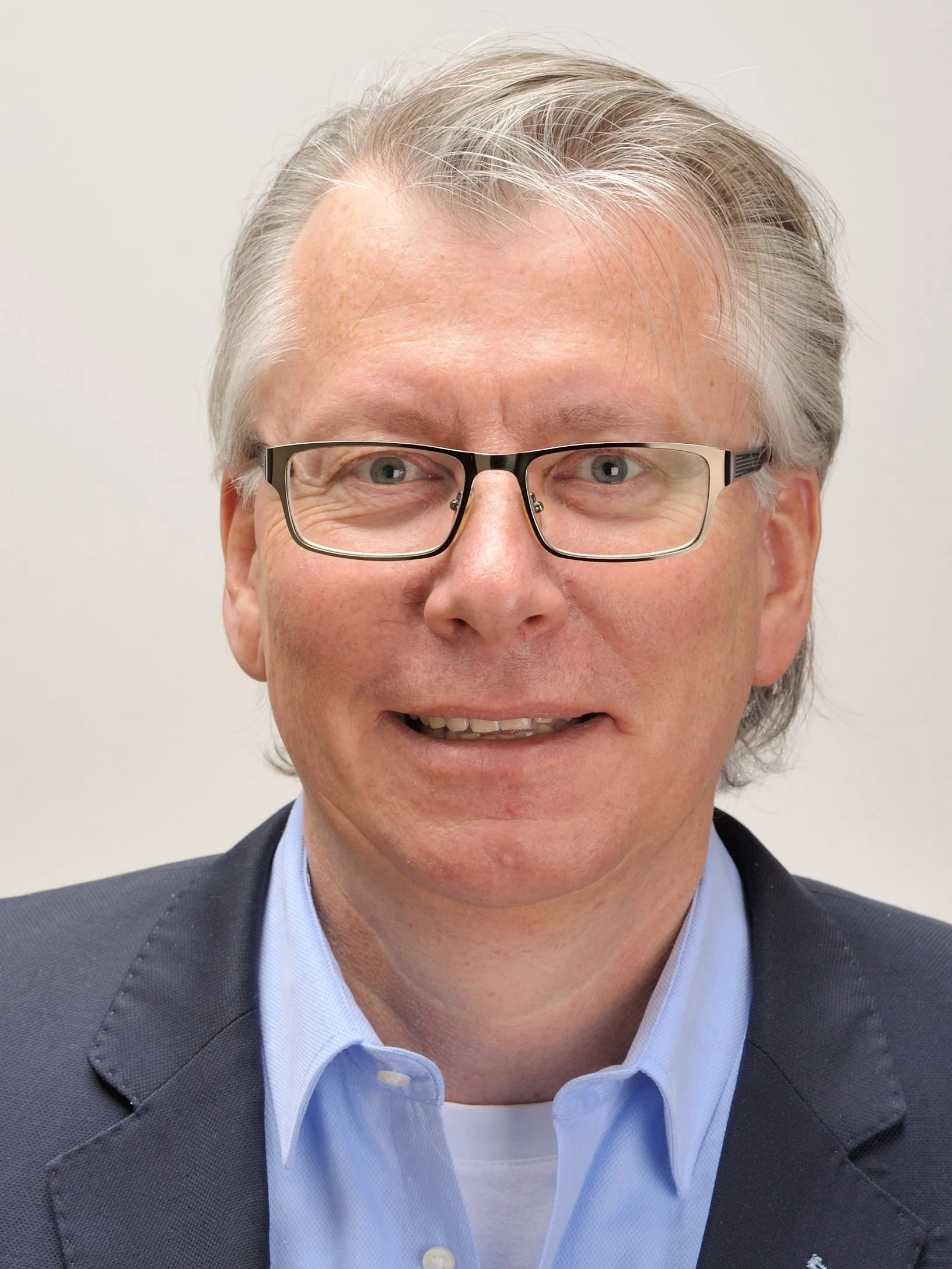 Ein Mann mit Brille in einem dunklen Blazer und blauer Hemd vor hellem Hintergrund, lächelnd.