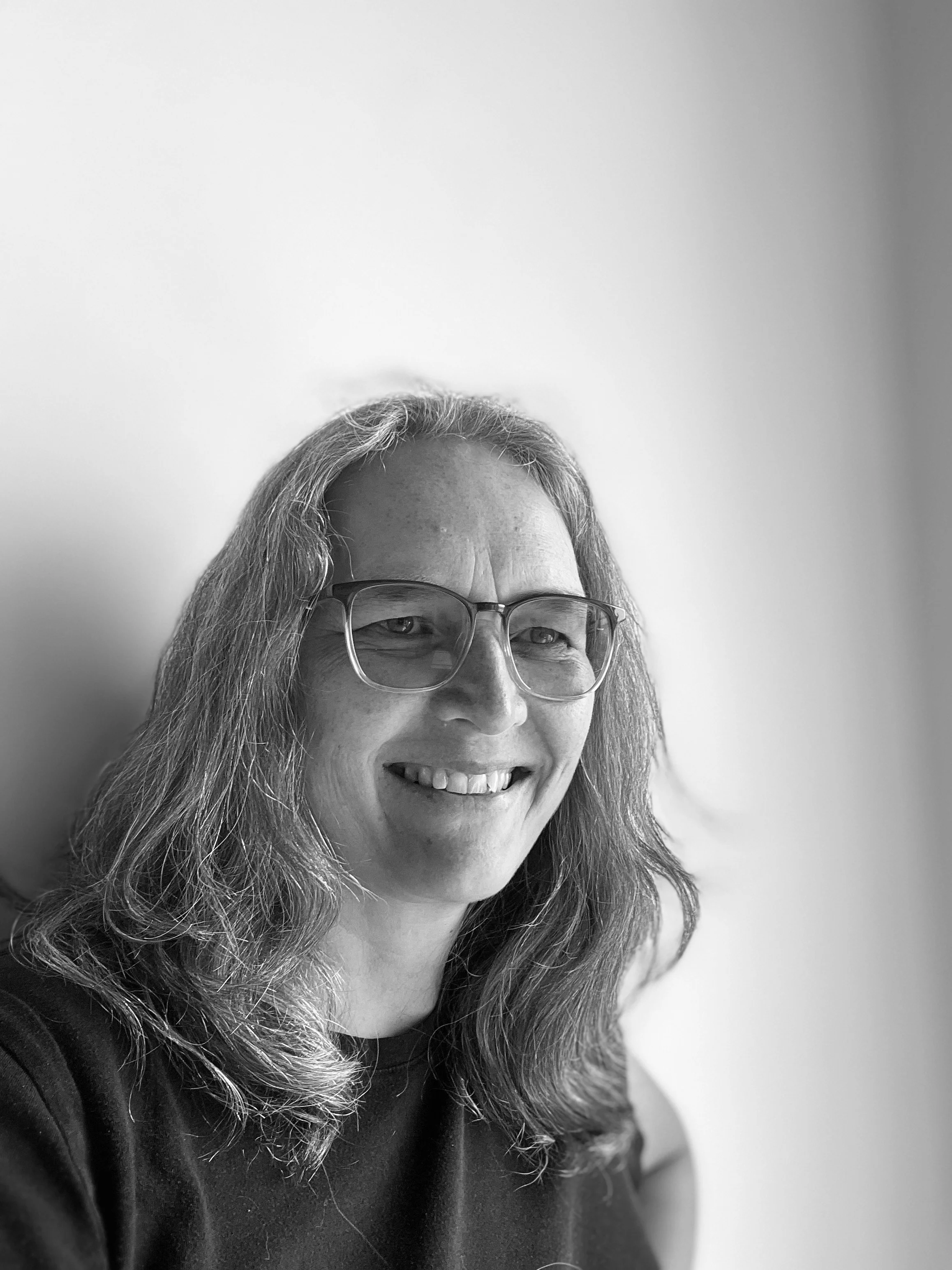 Black and white photo of a woman with wavy hair, wearing glasses, smiling, and seated against a plain background.