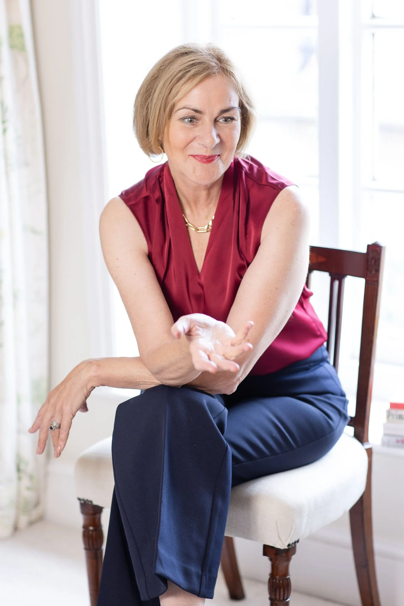 Maggie March sitting on a white cushioned chair, gesturing with her right hand while smiling slightly.