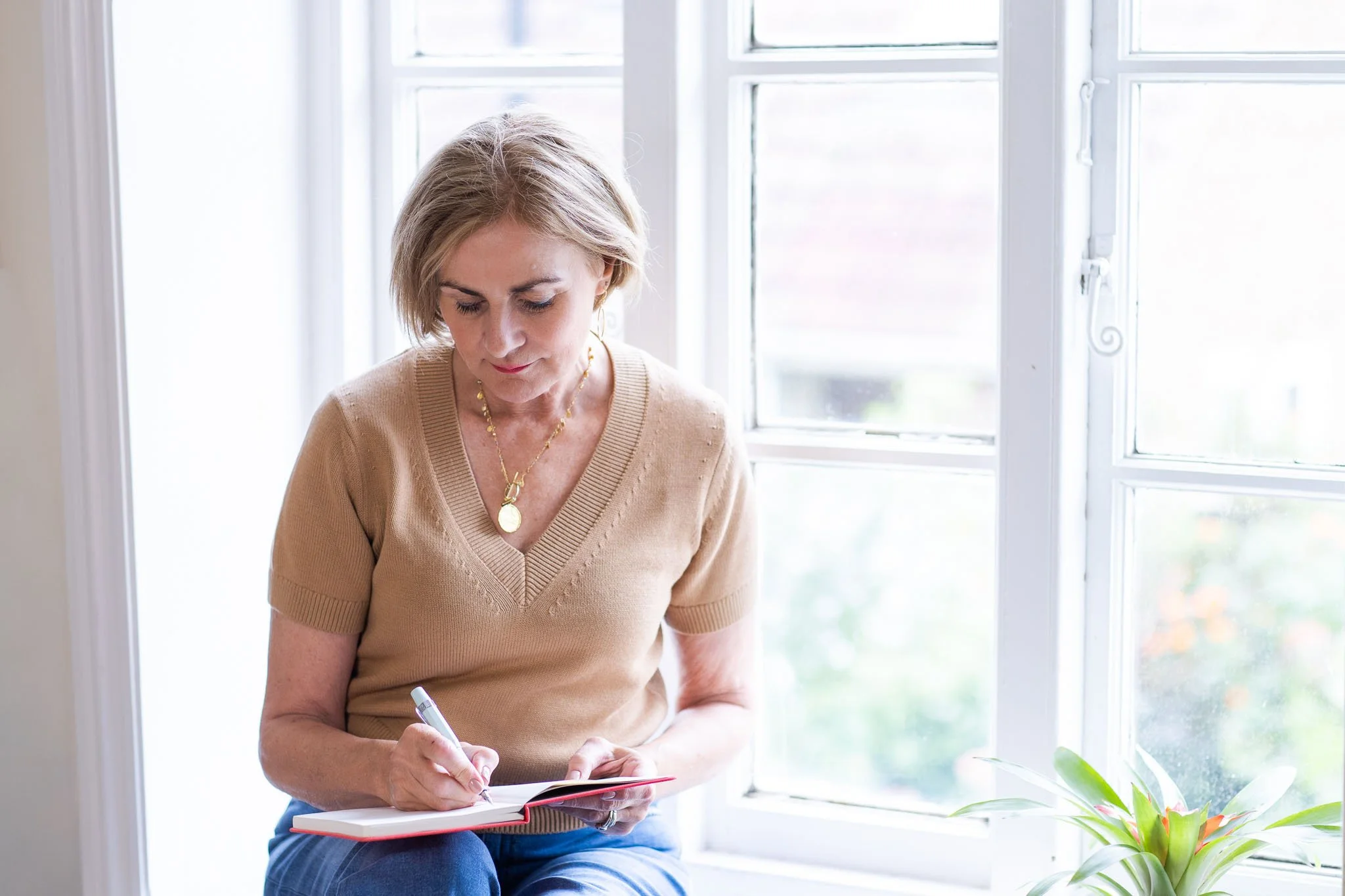 A woman sitting by a large window, looking down and writing in a notebook with a pen.