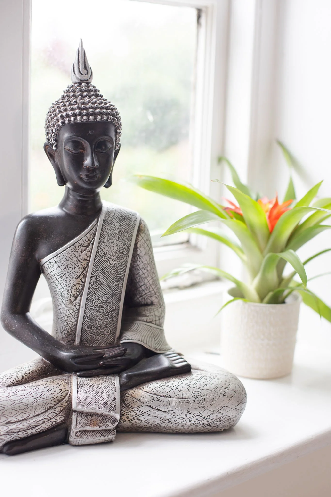 A black Buddha statue sitting in a lotus position next to a potted plant with green leaves and orange flowers on a white windowsill.