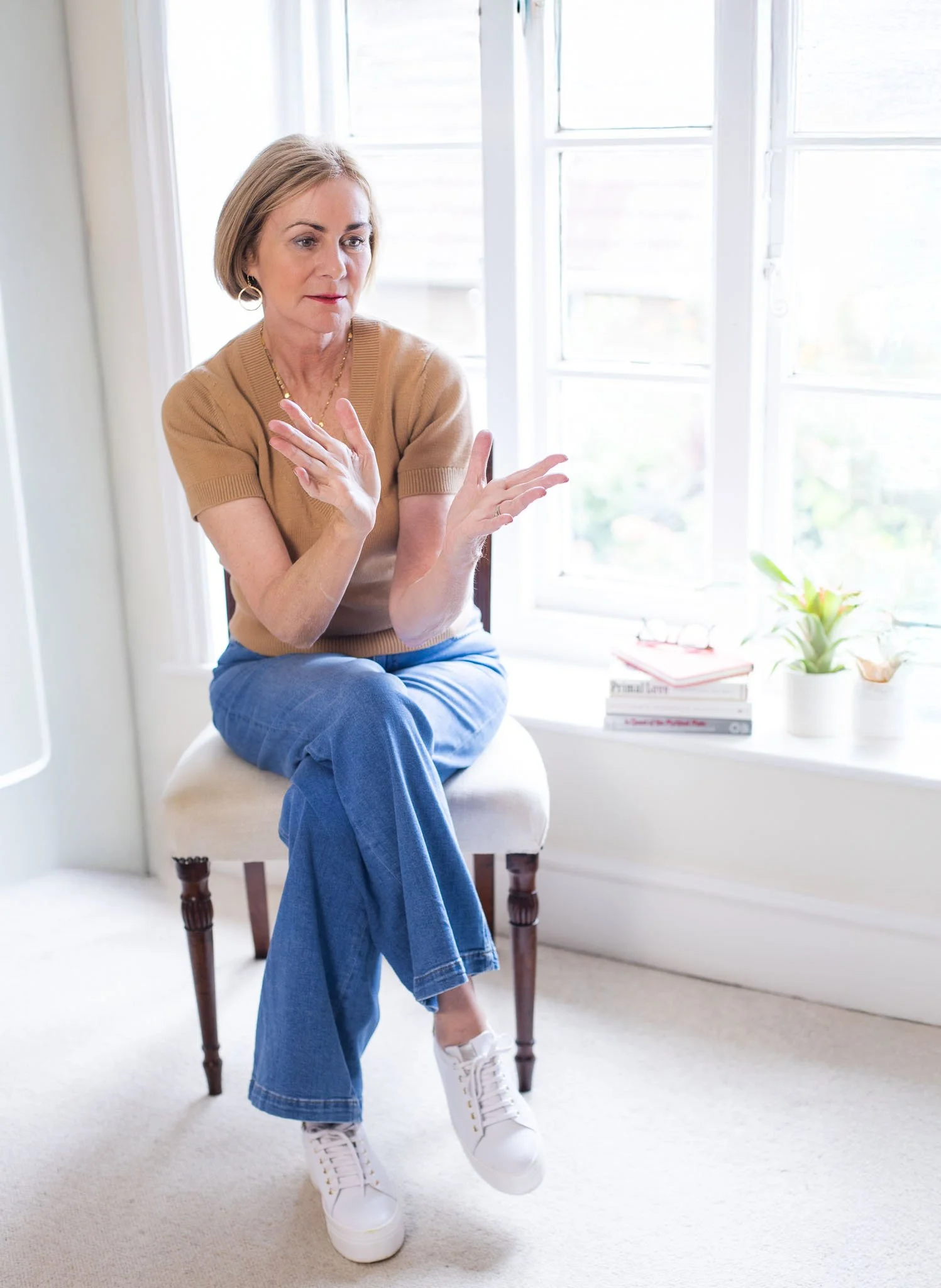 Maggie March sitting on a chair in a bright room near a large window, with books and a potted plant on the windowsill.