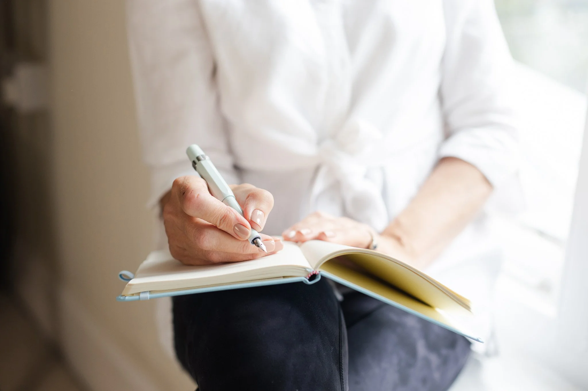 Maggie March writing in a notebook with a pen, sitting near a window.