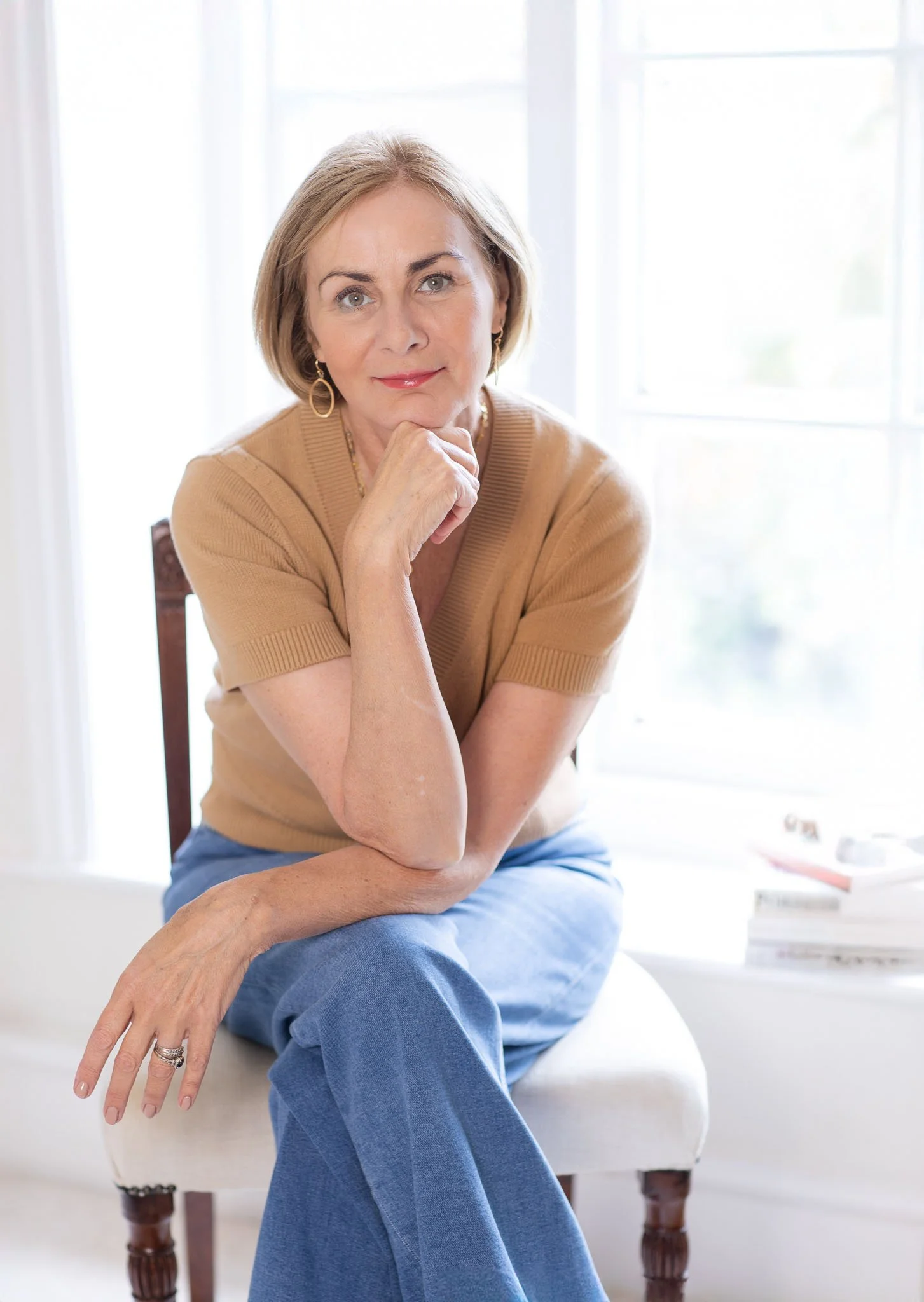 A woman with short blonde hair, wearing a tan short-sleeved sweater and blue pants, sitting on a wooden chair with a white cushion, resting her chin on her hand in front of a large window with natural light.
