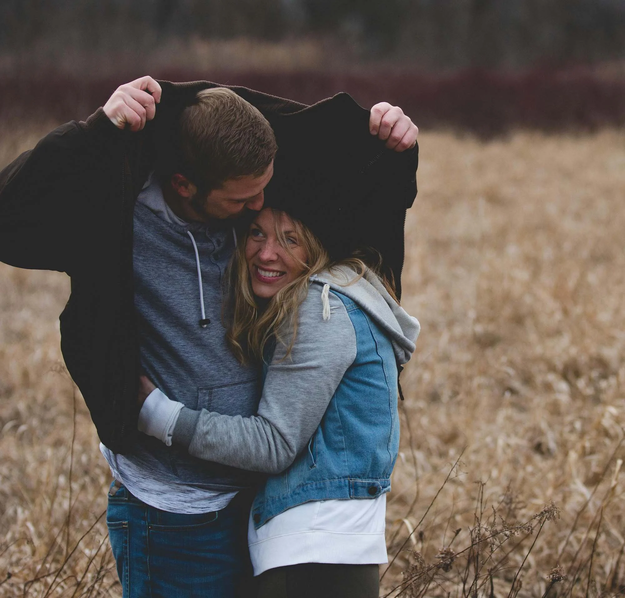A couple hugging outdoors in a field, with the man lifting the woman and the woman smiling.