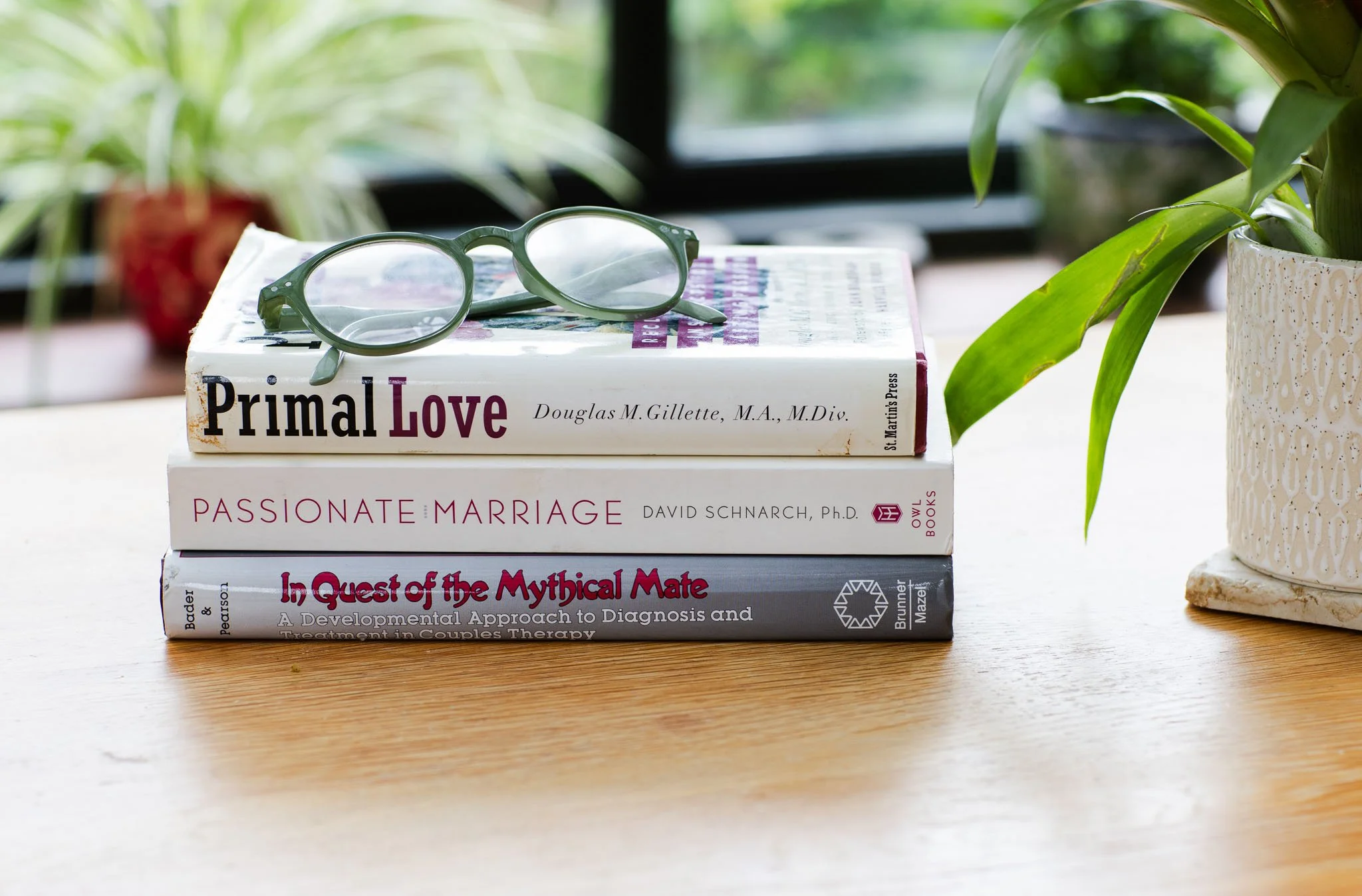 A stack of three books on a wooden table with glasses on top, surrounded by potted plants, near a window.