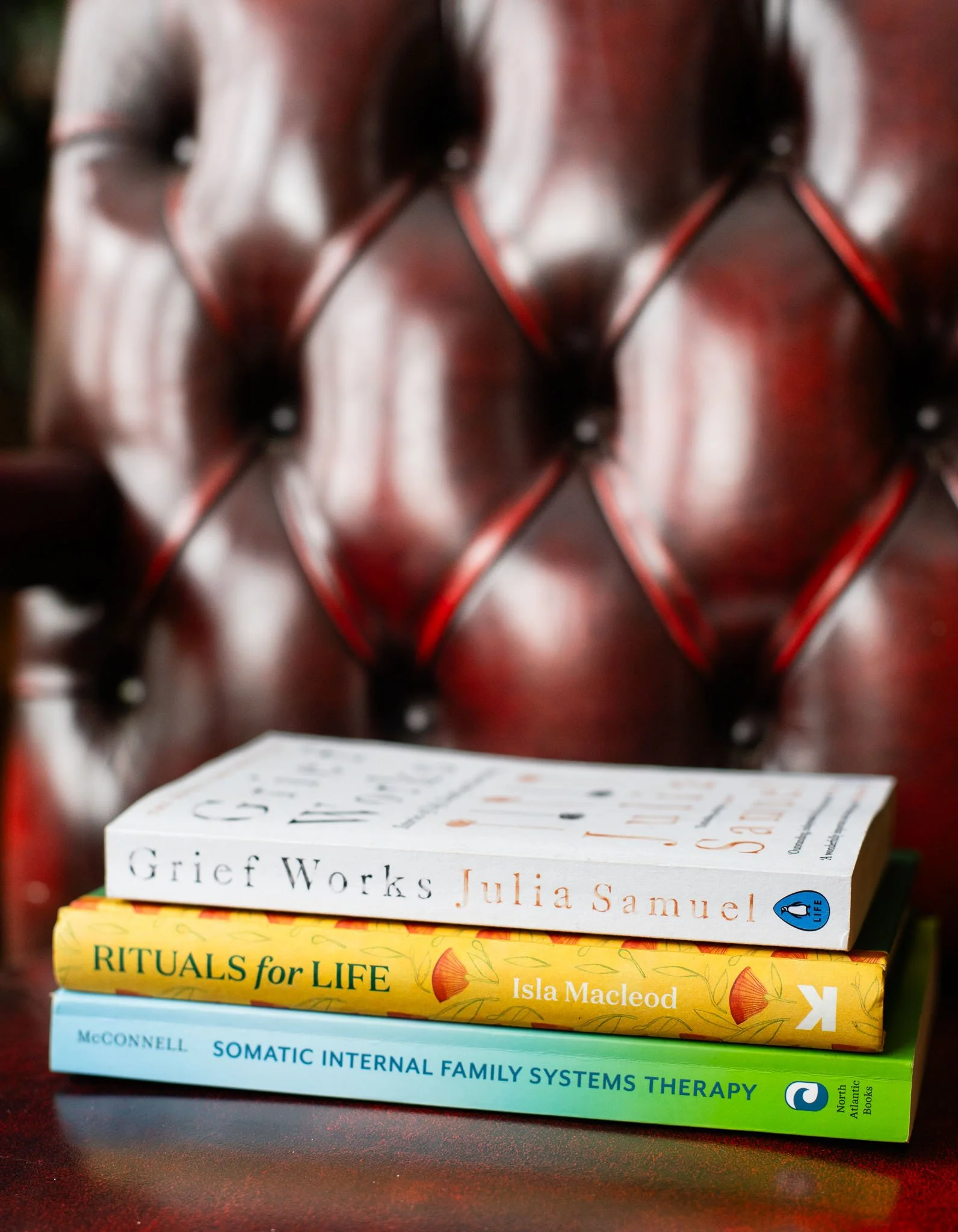 A stack of three books placed on a polished red leather tufted chair. The top book is titled 'Grief Works' by Julia Samuel. The middle book is titled 'Rituals for Life' by Isla Macleod. The bottom book is titled 'Somatic Internal Family Systems Therapy' by McConnell.