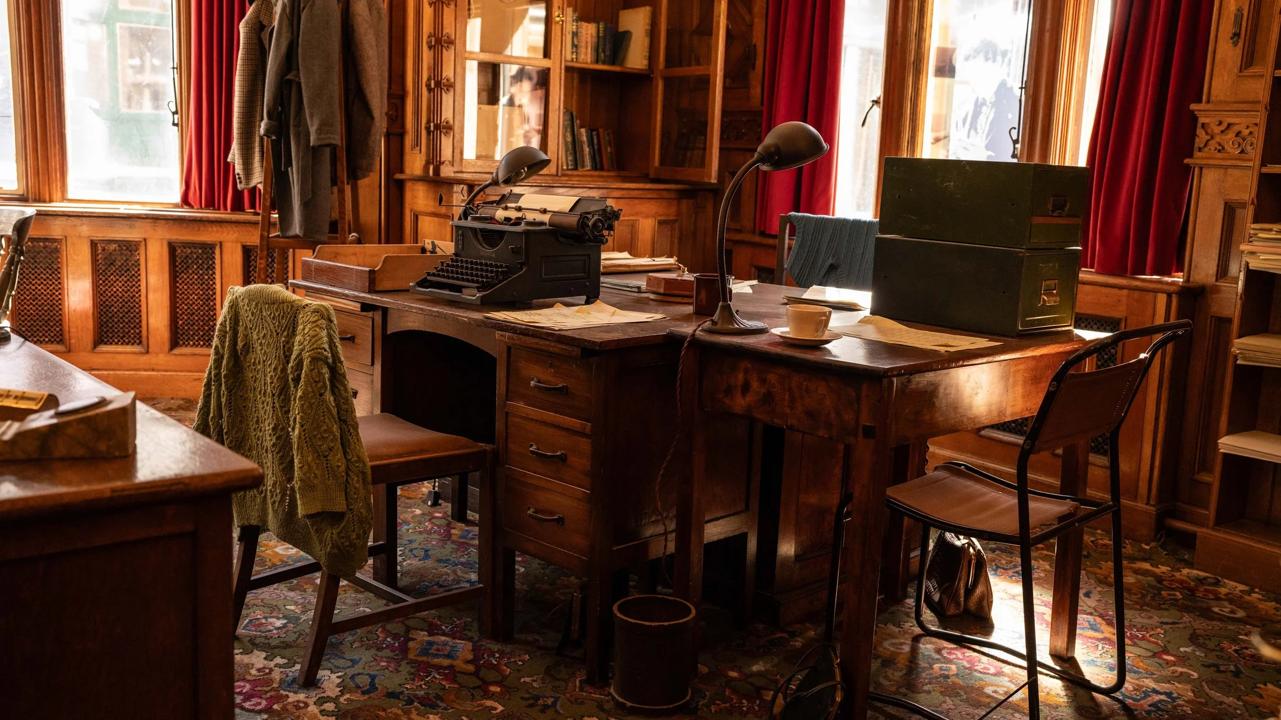 A vintage-style wooden office with two desks, one with an old typewriter and the other with files and papers, surrounded by chairs, bookshelves, and windows with red curtains.