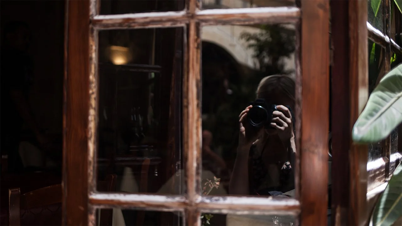 A person taking a photo of their reflection in a glass window or mirror, framed by wooden window panes, with some greenery visible on the right side.