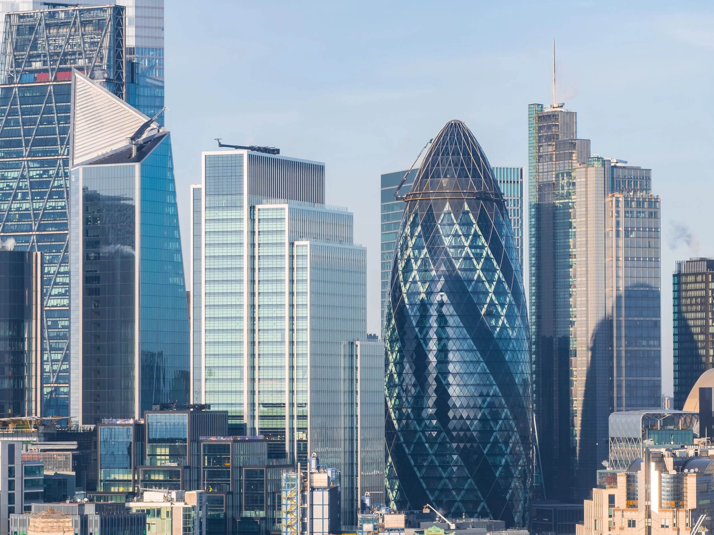Skyline of modern skyscrapers including the Gherkin building in London with distinctive oval shape and glass exterior, glass office towers, and city rooftops under a light blue sky.