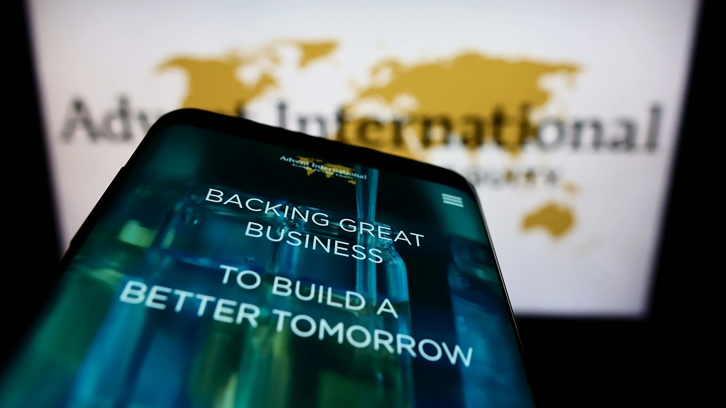 Close-up of a mobile phone displaying a website with the text "Backing Great Business To Build A Better Tomorrow". In the background, a blurred screen shows the logo of Australian International.