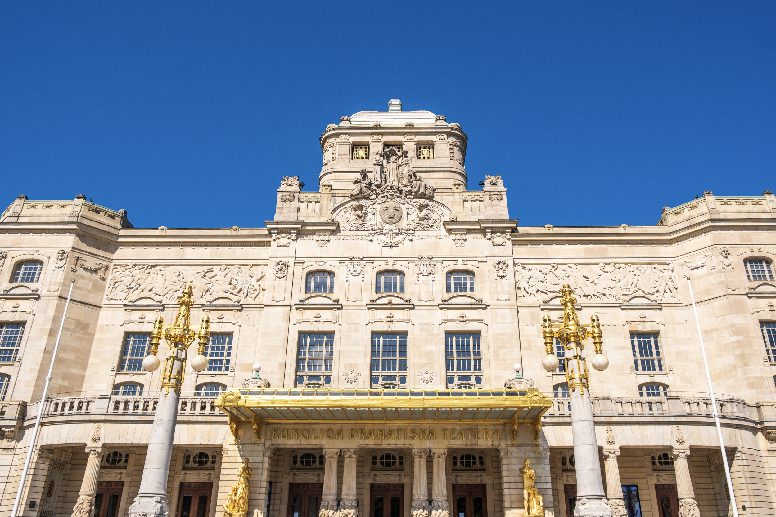 Facade of the Kungliga Dramatiska Teatern building with detailed sculptures, columns, and golden accents, under a clear blue sky.