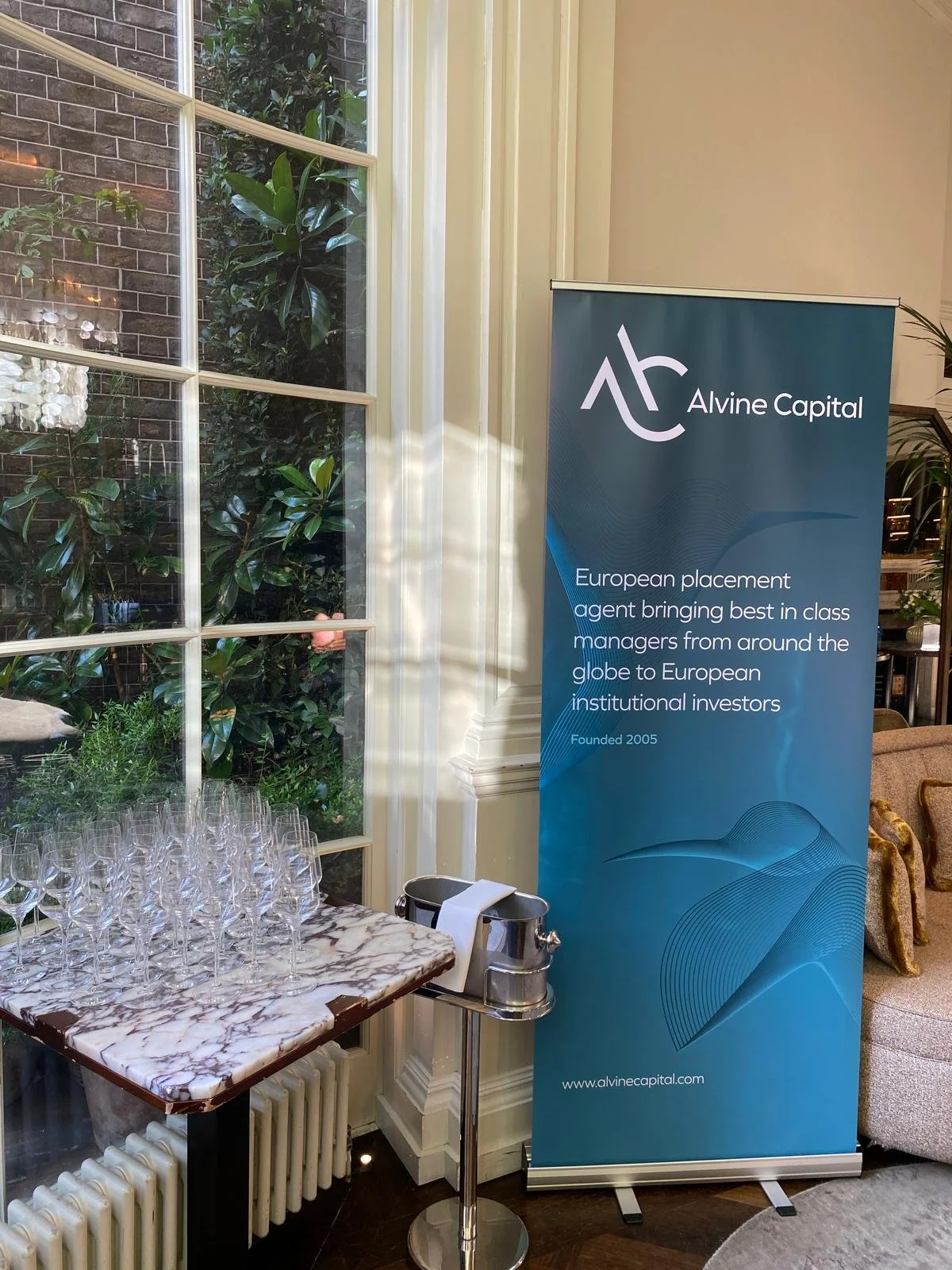 A blue standing banner with Alvine Capital logo and text about European placement agents, next to a table with empty wine glasses and a window with greenery outside.