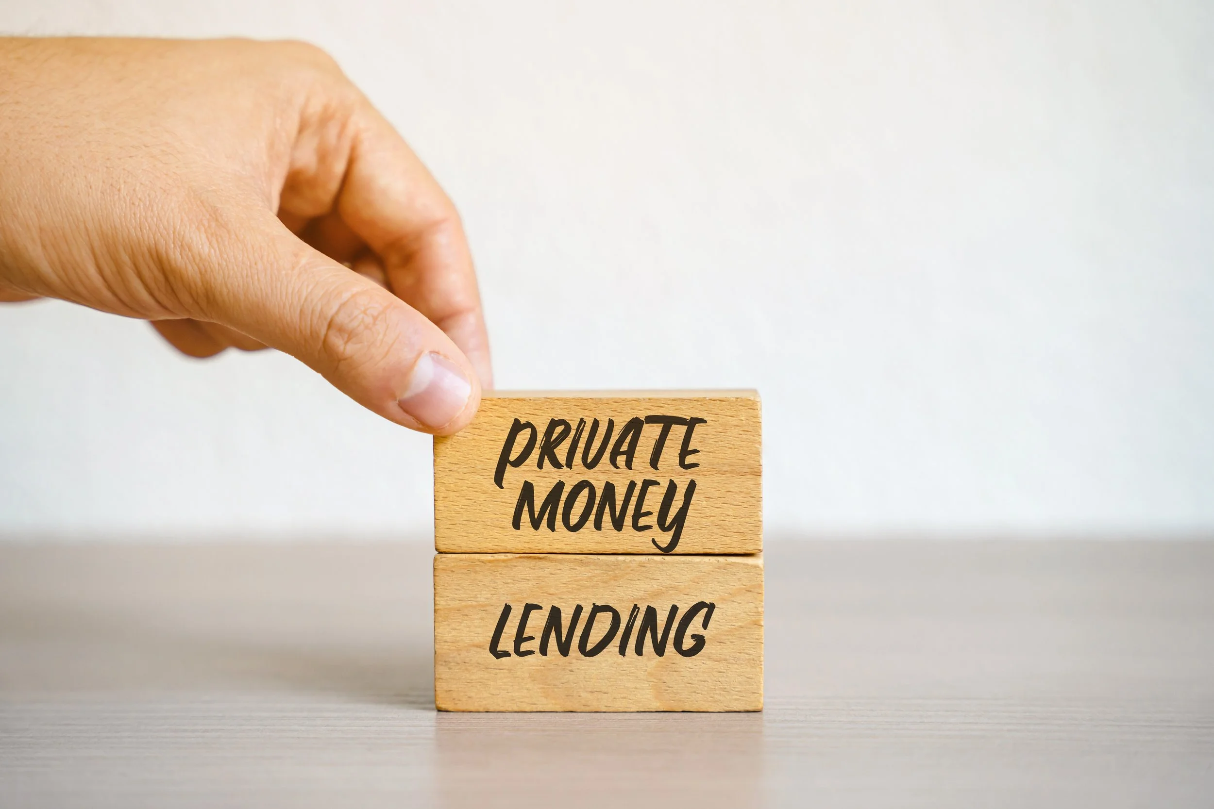 Close-up of a hand stacking two wooden blocks with the words 'private money' on the top block and 'lending' on the bottom block on a light-colored surface.