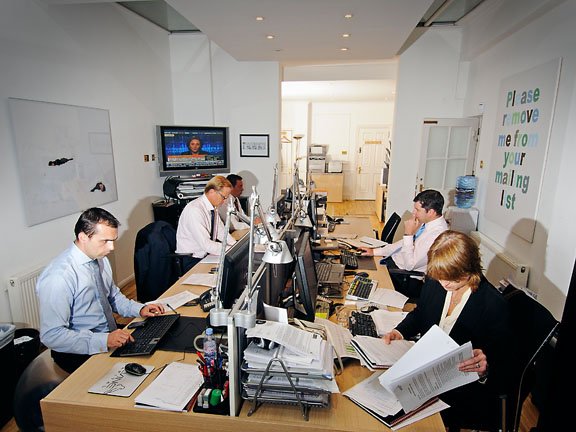 Office scene with five people working at desks with computers and papers.