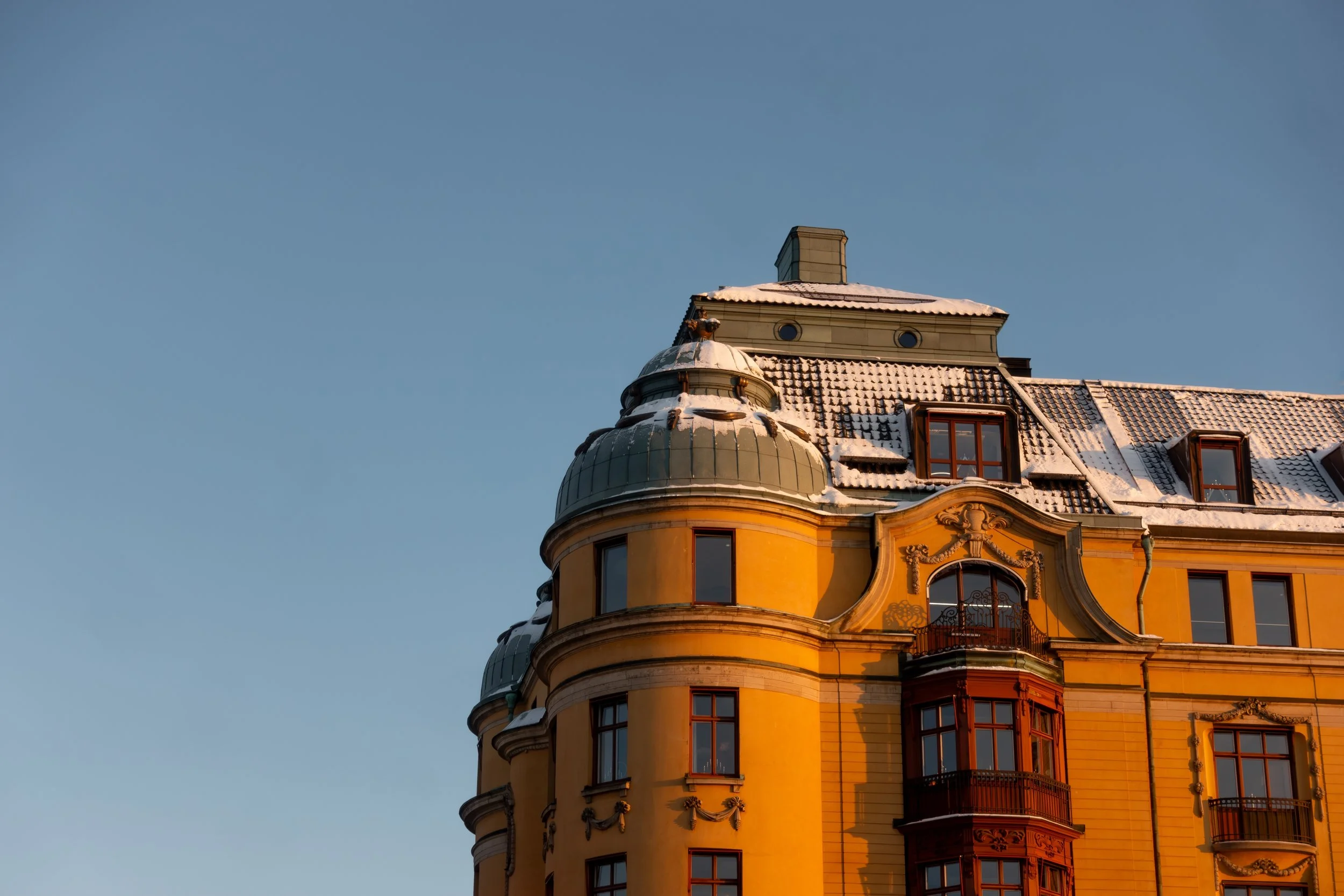 Close-up of a yellow historic building with ornate architectural details, balconies, and a rounded tower, topped with snow, against a clear blue sky.