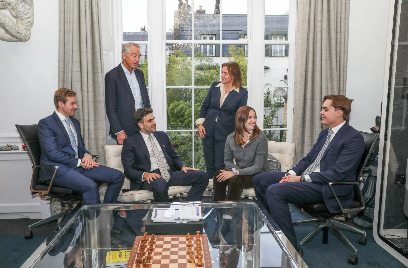 Business meeting in a bright office with six people, five sitting and one standing, having a discussion, with a chessboard on a glass table in front.