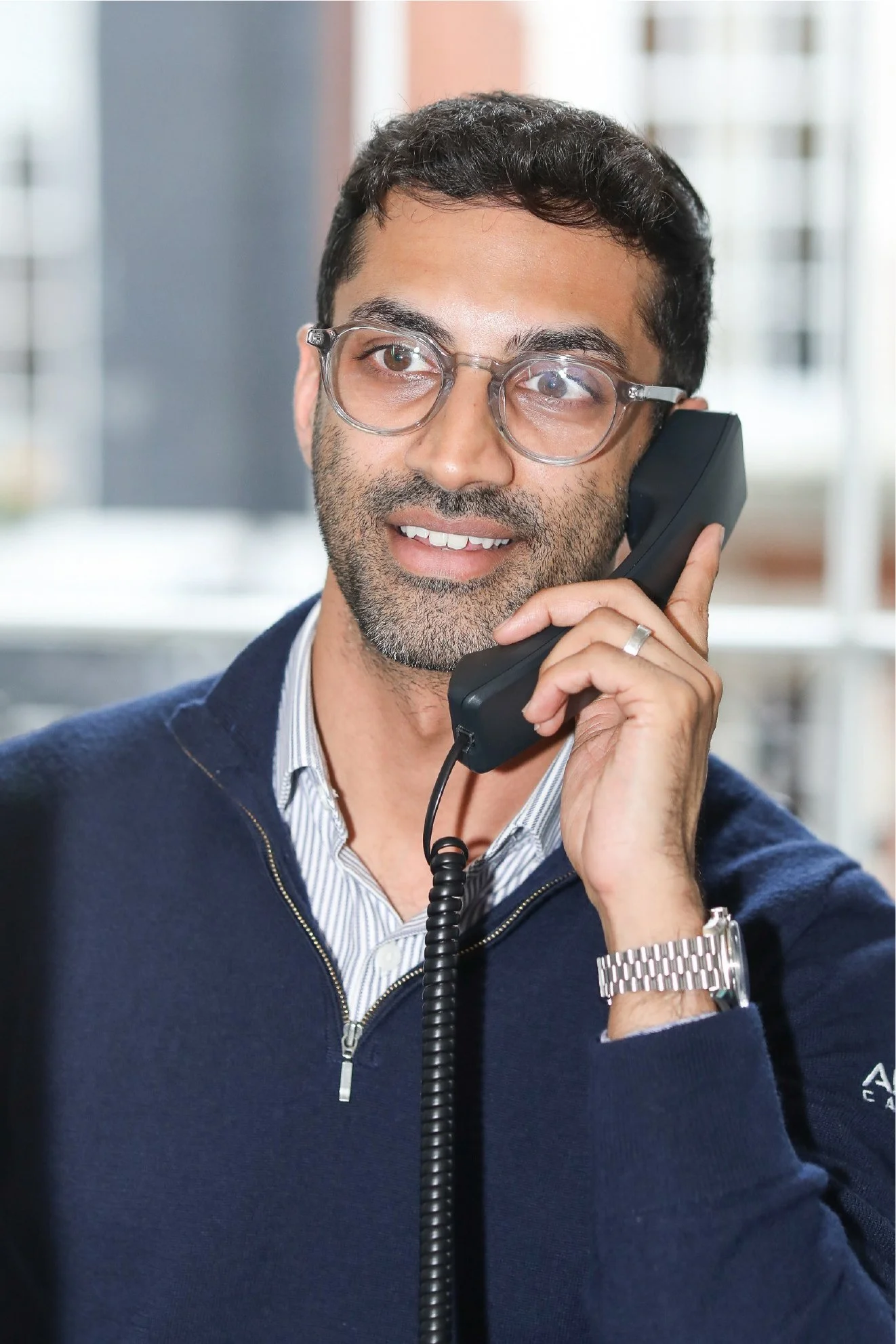 A man with dark hair, beard, and glasses, wearing a dark blue zip-up sweater, is talking on a corded telephone in an office environment.