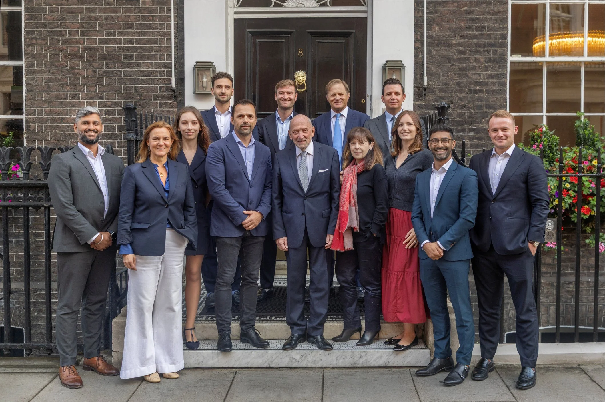 A group of fifteen professionally dressed people standing on the steps of a brick building with a black door, posing for a photo outdoors.