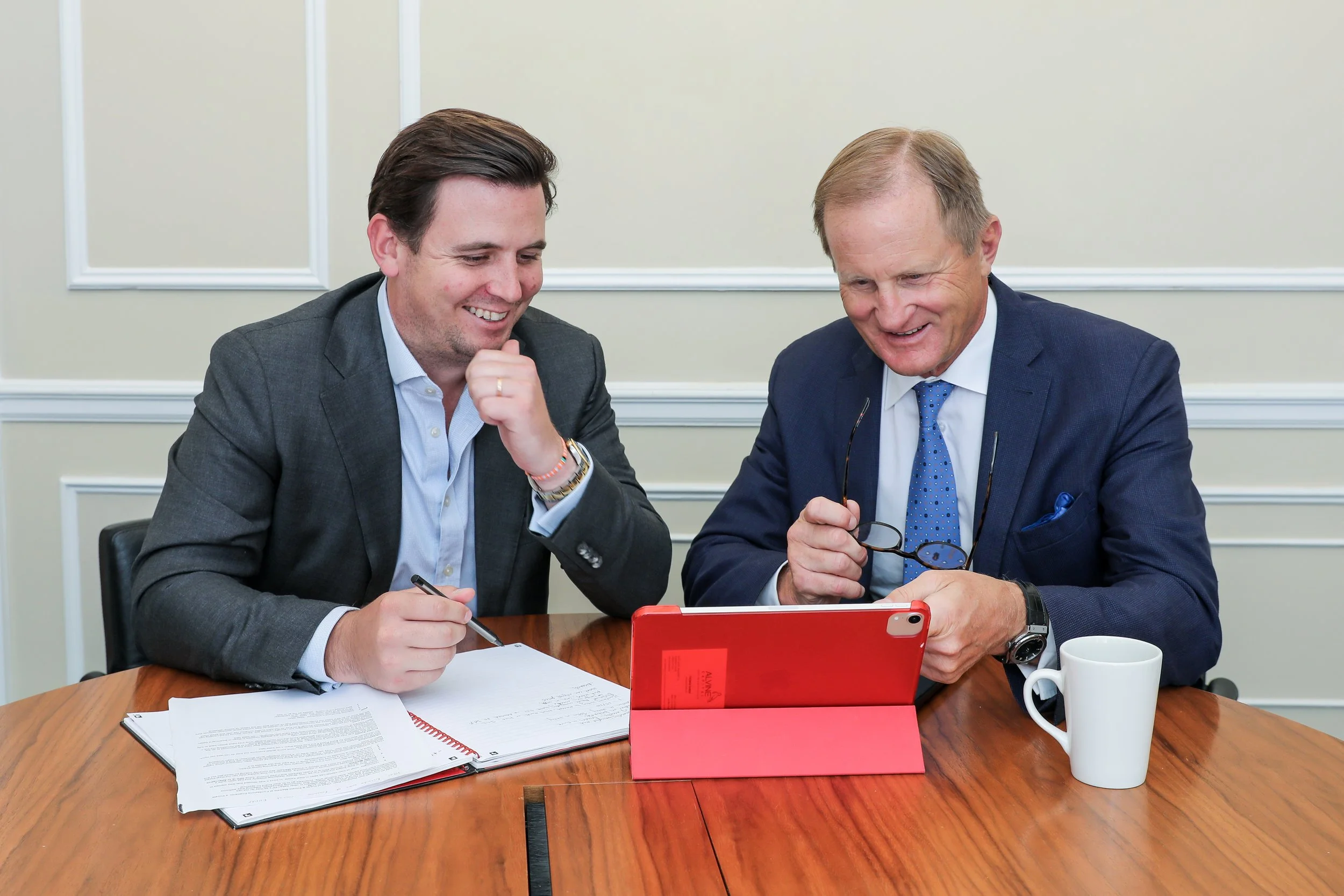 Two men in business suits sitting at a table, looking at a tablet and smiling, with papers and a mug on the table.