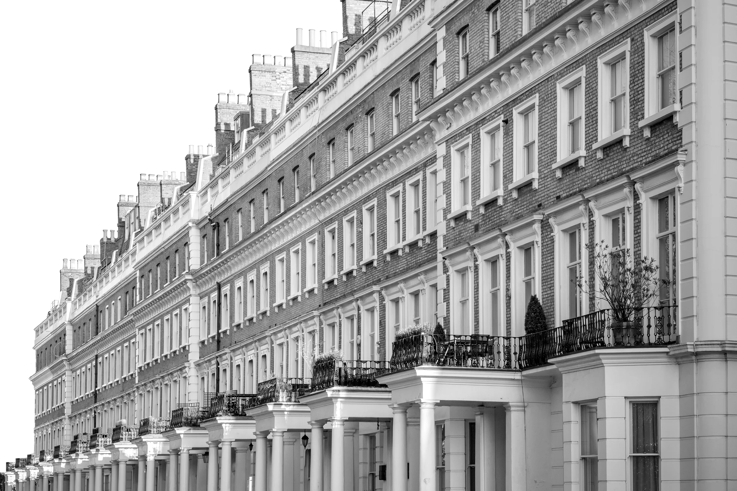 Black and white photo of a row of brick residential buildings with multiple stories, balconies, and large windows, featuring classical architectural details such as columns and decorative cornices.