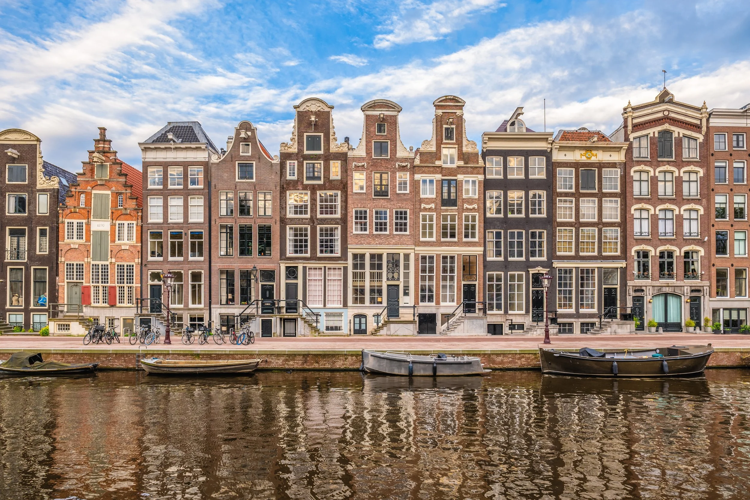Colorful row of historic European-style townhouses along a canal with boats and bicycles parked on the sidewalk, under a partly cloudy sky.