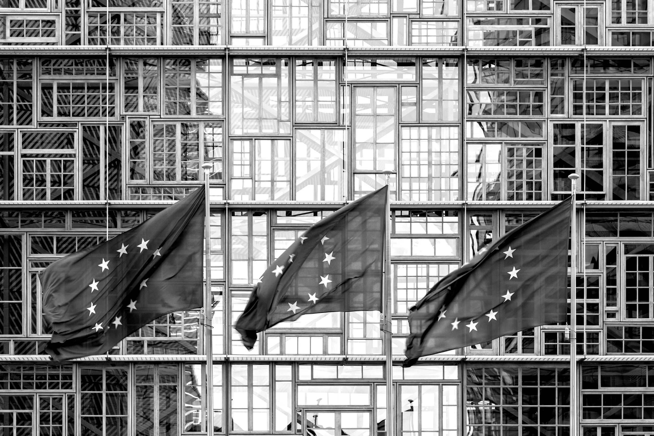 Three European Union flags flying in front of a large modern glass building.