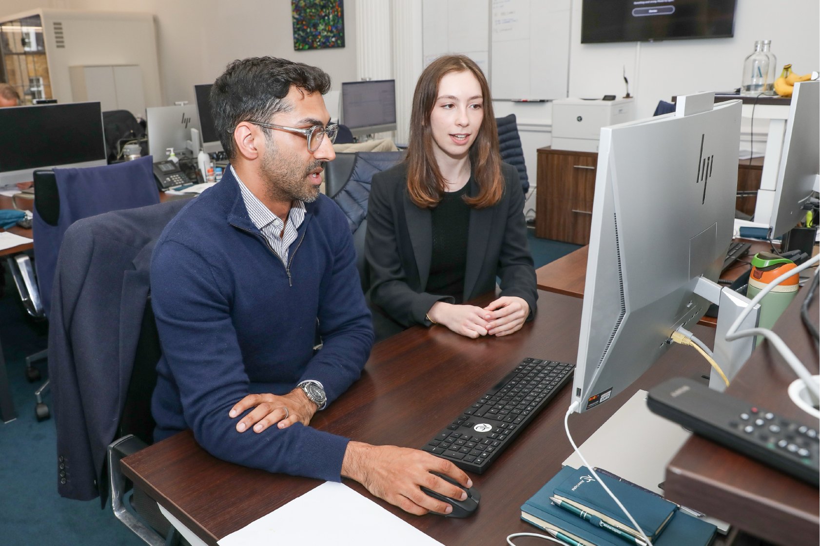 Two professionals, a man and a woman, working at a desk with computers in an office setting.