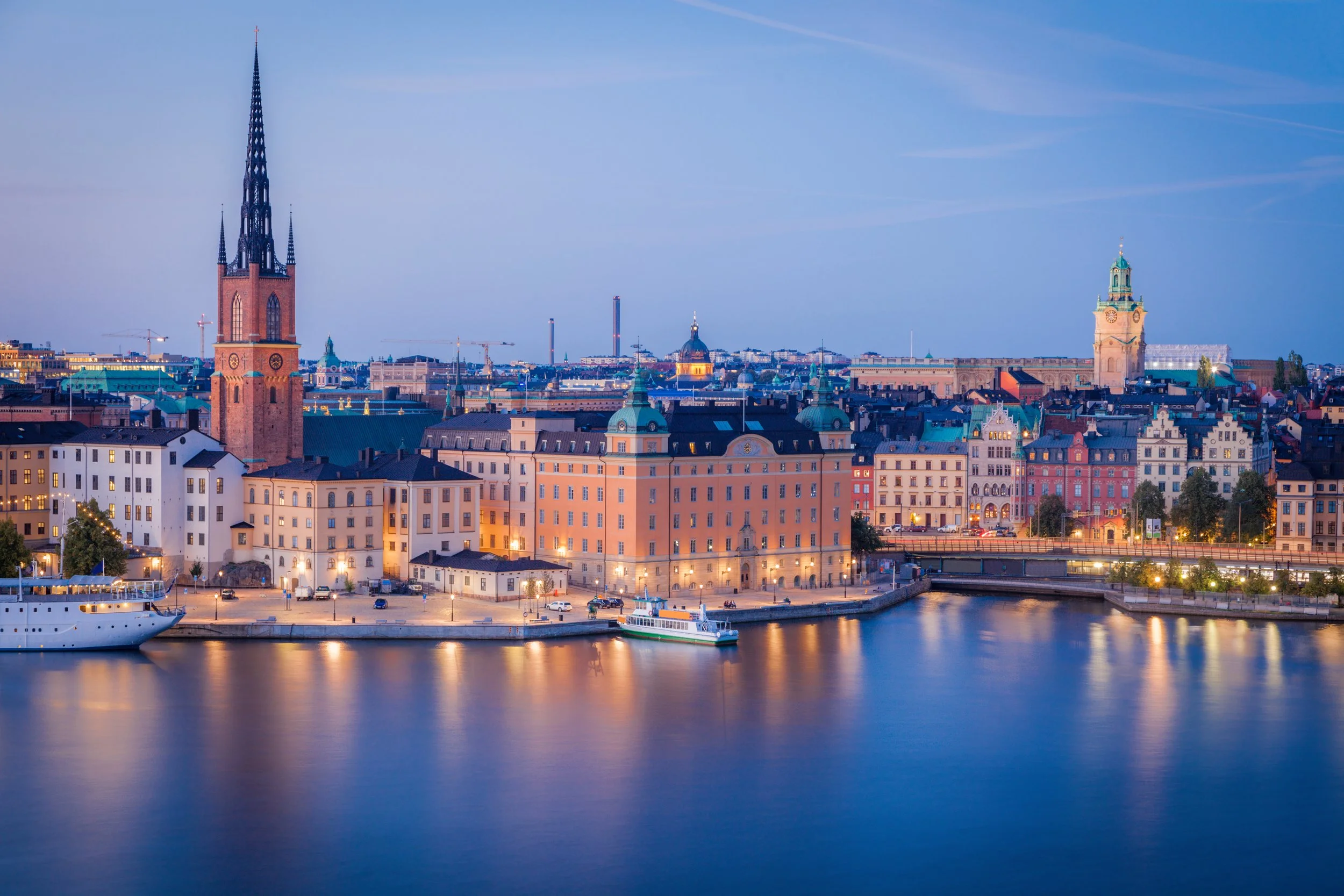Panoramic view of Stockholm, Sweden at dusk featuring historic buildings along the water, including a prominent church with a tall spire, and calm water reflecting city lights.