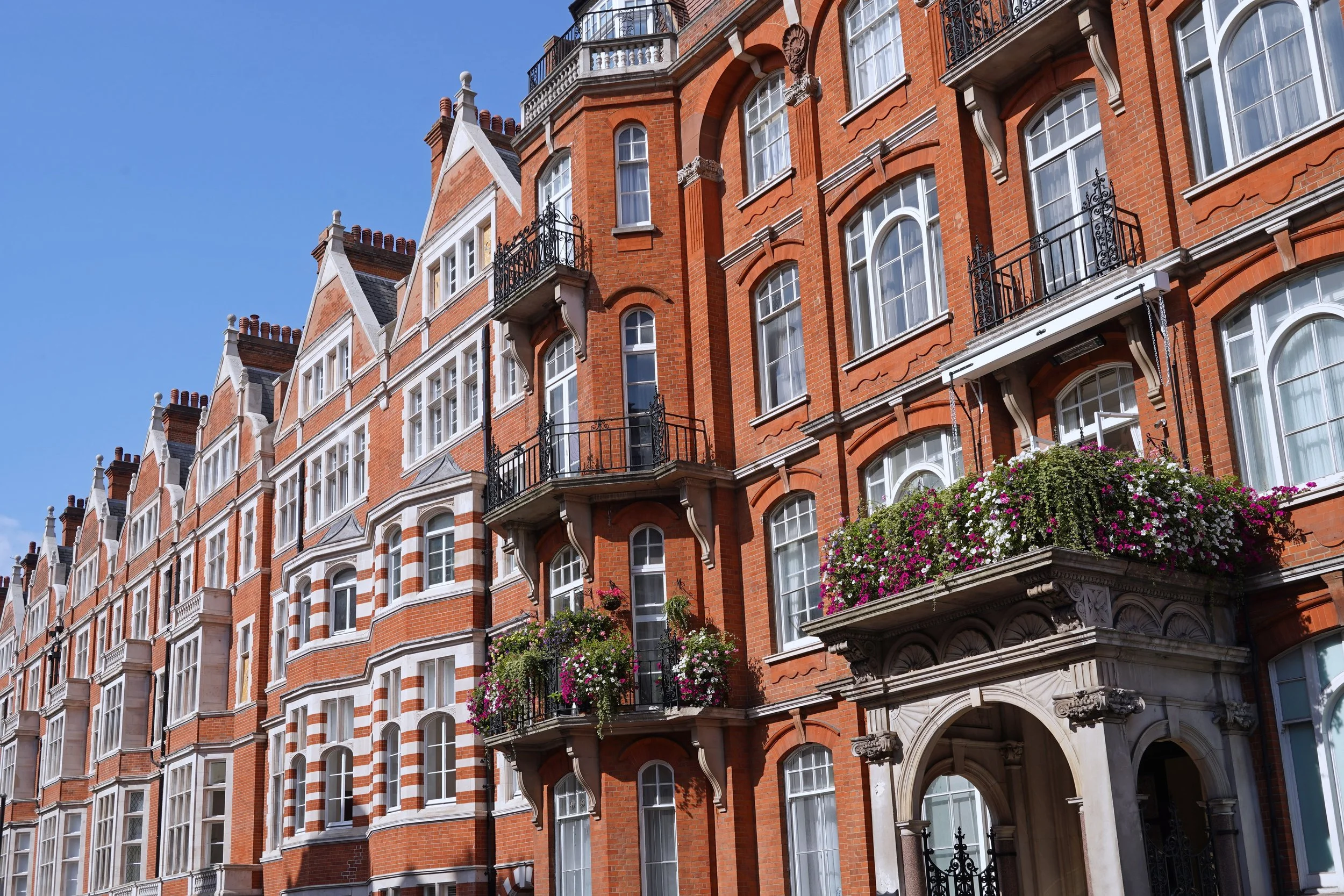 A large, multi-story red brick apartment building with white window frames and balconies decorated with hanging flower baskets, set against a clear blue sky.