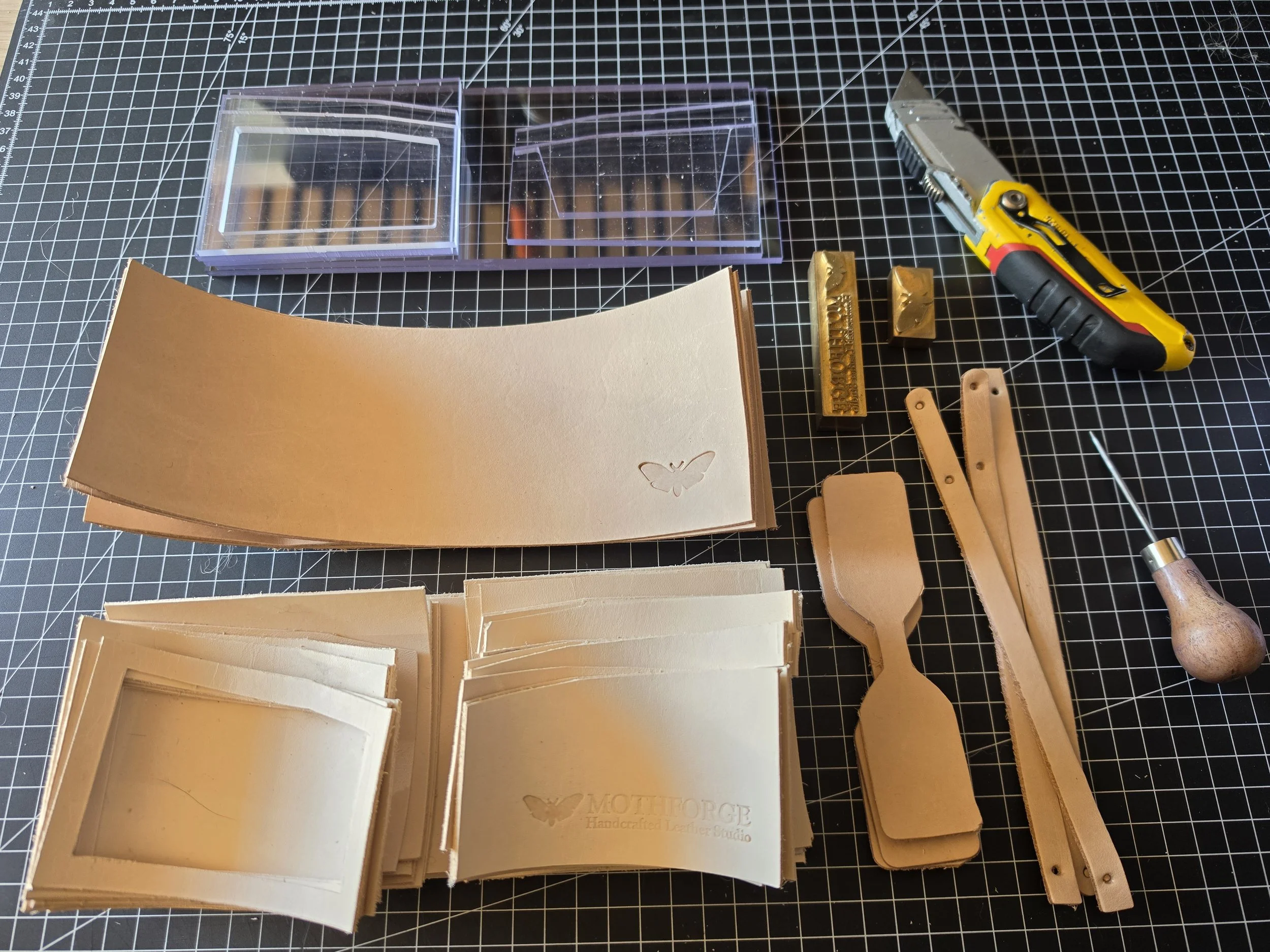 Collection of crafting supplies on a grid cutting mat, including clear plastic boxes, leather pieces, metal tools, and a utility knife.